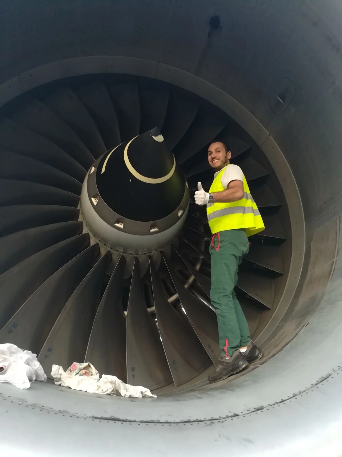 Uomo con giacca gialla ad alta visibilità e pantaloni verdi sorridente e che fa il gesto del pollice in su accanto a una turbina eolica, all'interno di un tunnel della turbina.