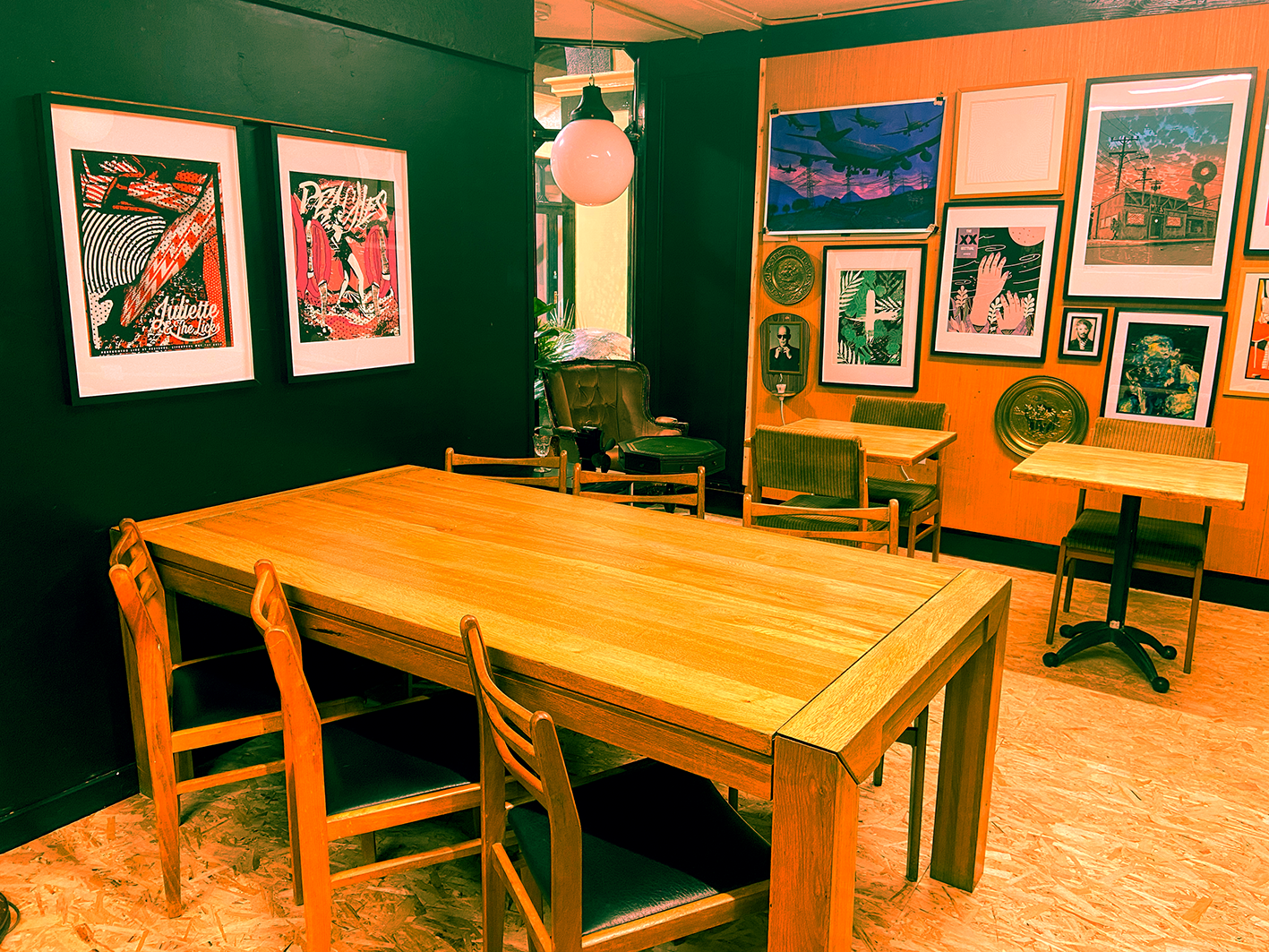 Interior of a cozy room with a large wooden table, surrounded by mid century wooden, adorned with colorful artwork and photographs on the walls, and illuminated by a round hanging light.