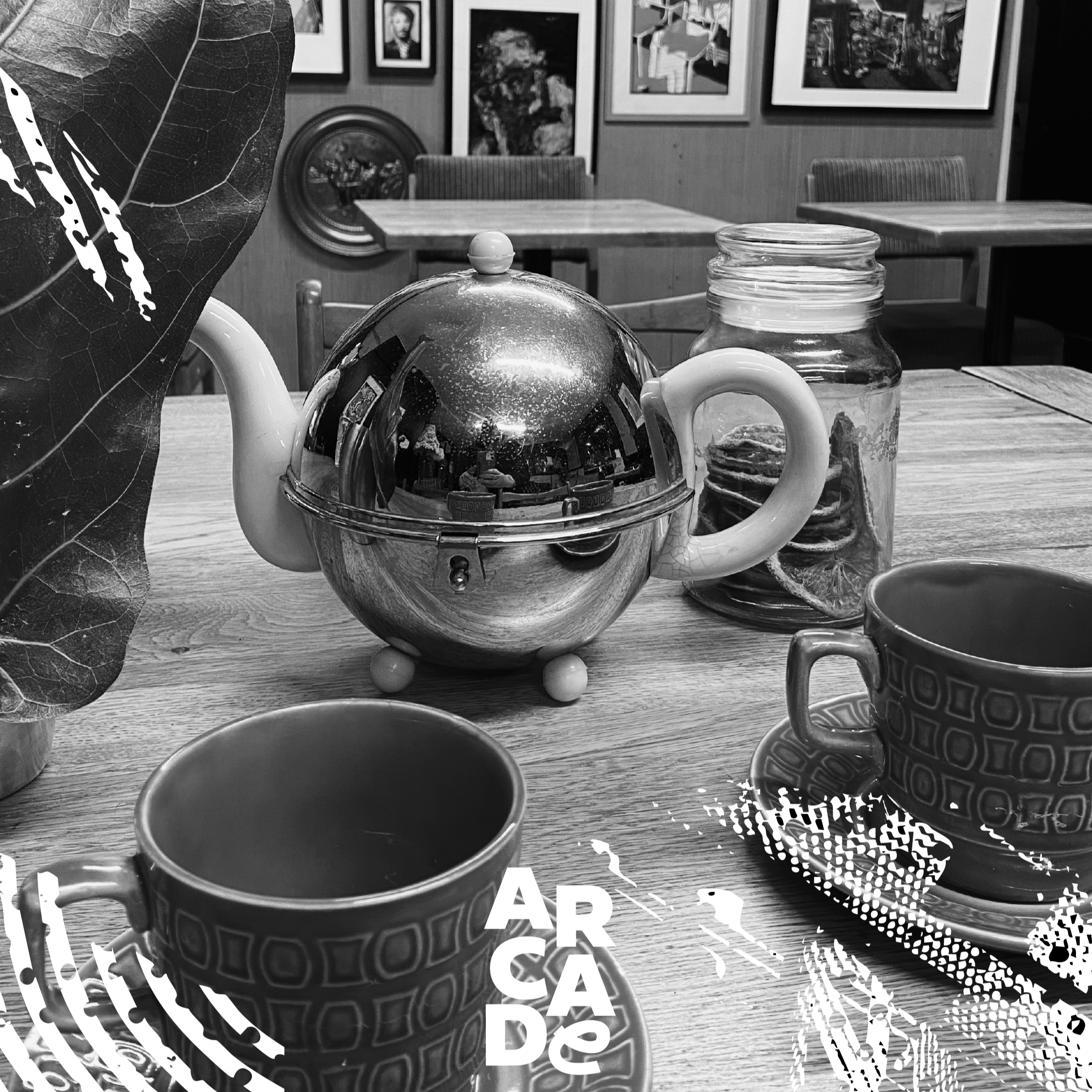 Black and white photo of a wooden dining table with a shiny teapot, a jar with cookies, two patterned cups on saucers, and a leafy plant in a pot. The background shows framed pictures on the wall and a couple of chairs.