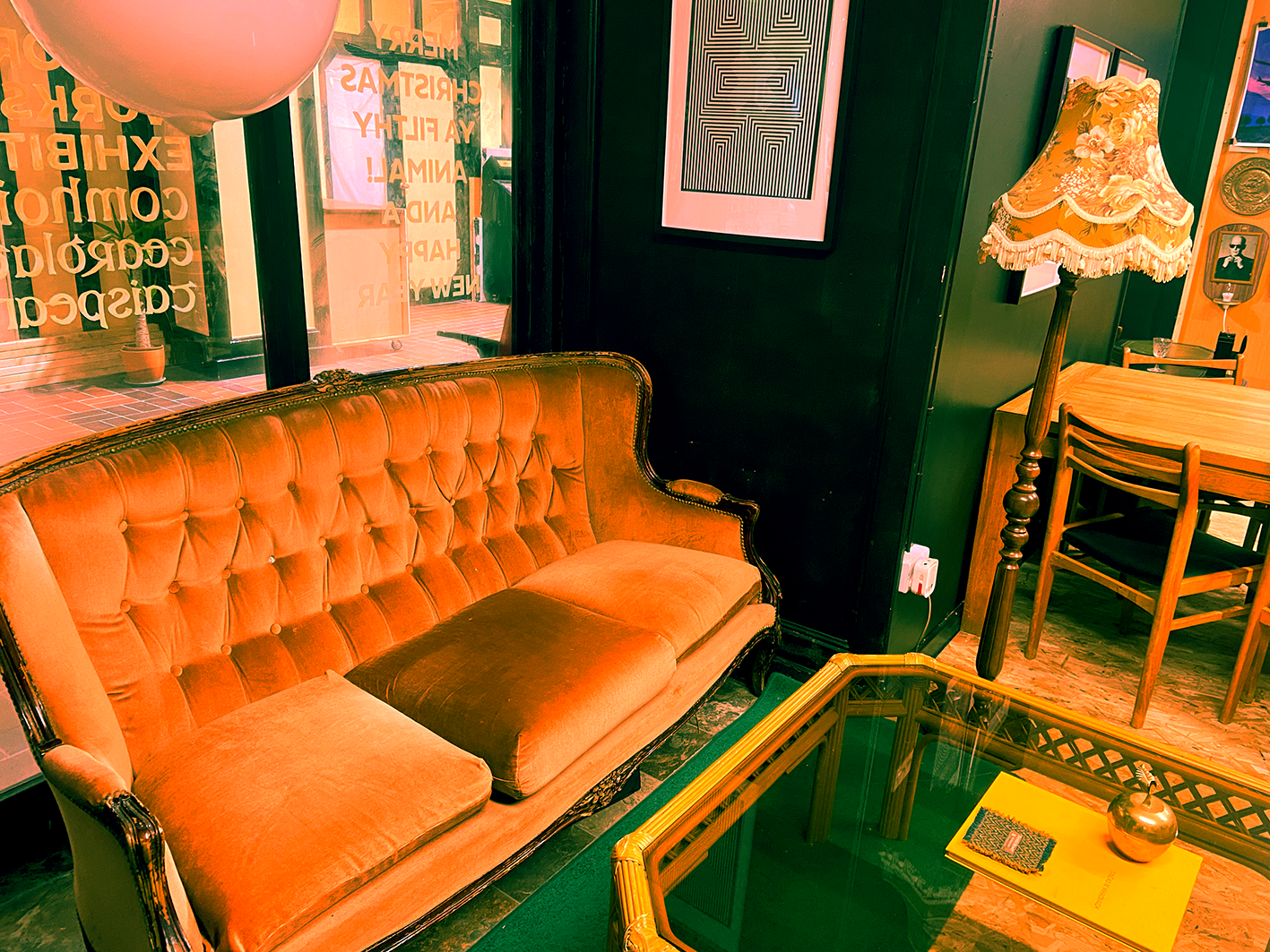 Vintage style interior with an orange velvet tufted sofa, a tall floor lamp with a floral patterned shade, a wooden dining table with chairs, artwork on the dark green walls, and a window with a sign in the background.