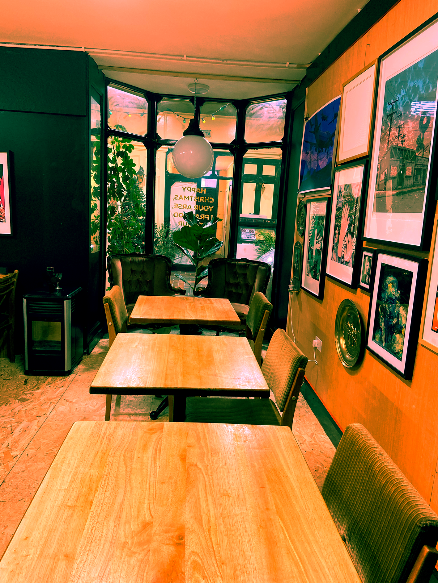 Interior of a cozy cafe with wooden tables and chairs, framed artwork on the wall, a green plant near the window, and a large spherical hanging light.