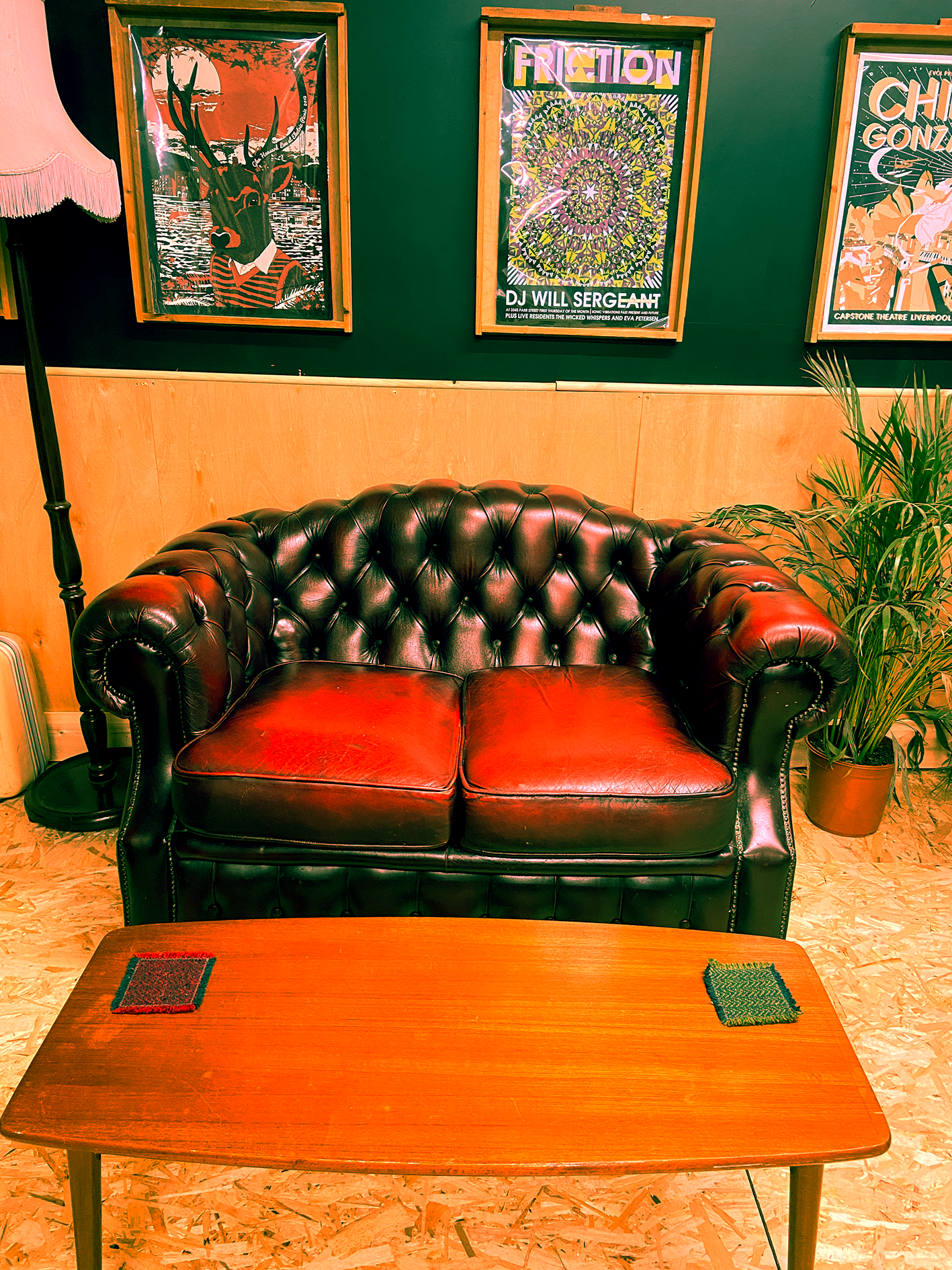A vintage leather tufted sofa with a mid century wooden coffee table in front of it, positioned in a cozy room with posters on the wall and a potted plant on the right. The posters feature colorful and artistic illustrations.