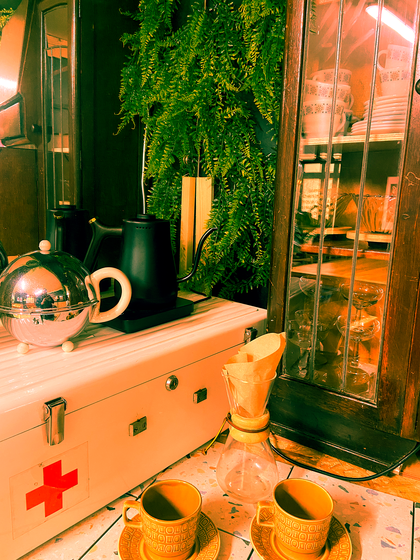A cozy kitchen corner with a vintage white cabinet featuring a red cross symbol, a glass coffee pour-over setup, two black electric kettles, a large green leafy fern plant, and a glass vase with a paper-filtered coffee dripper. Two yellow patterned cups with matching saucers are in the foreground on a tiled floor, and a glass cabinet with wooden framing displays dishware inside.