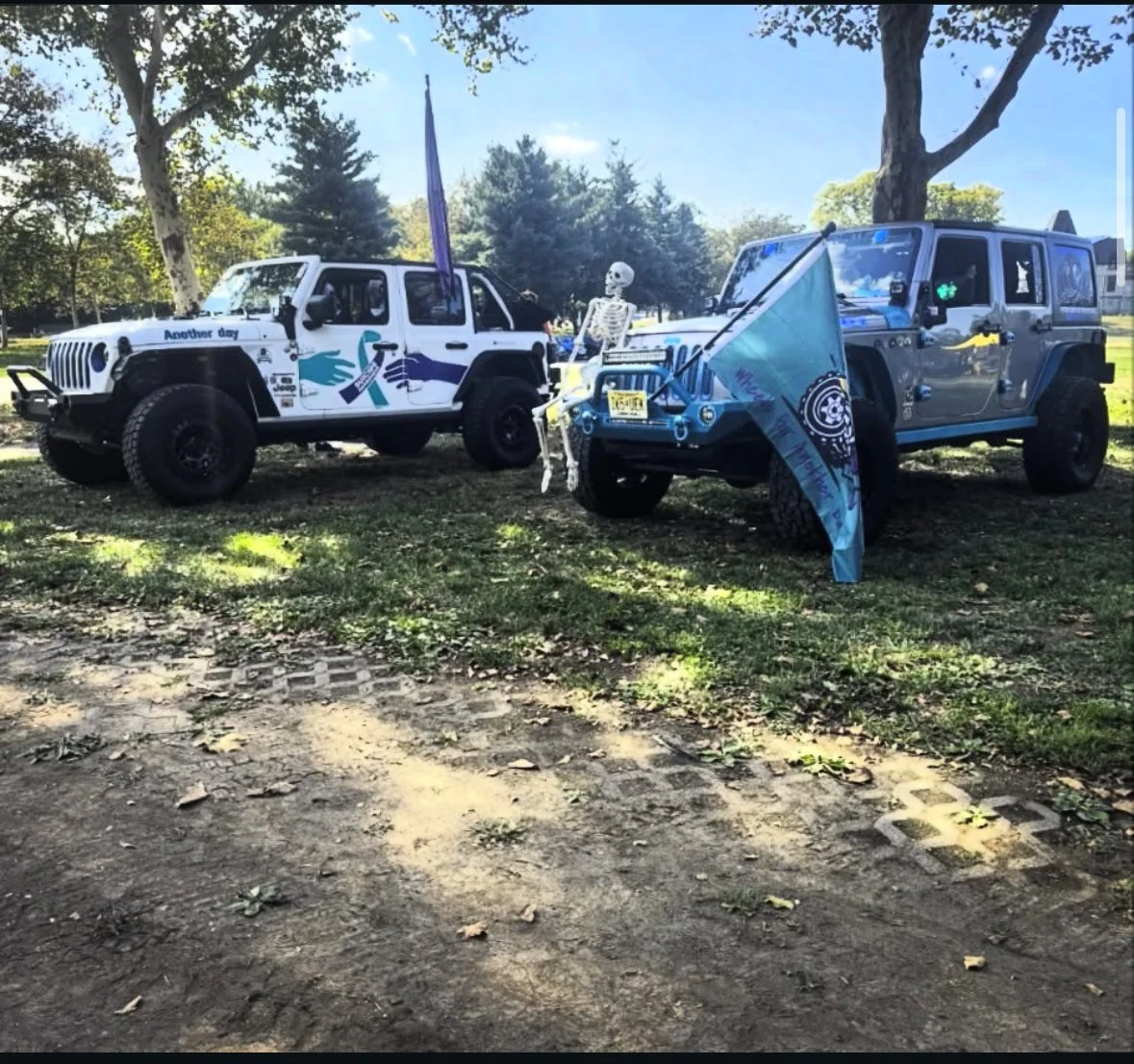 Two decorated Jeep vehicles parked outdoors with a skeleton figure between them, holding a flag, under trees on a sunny day.