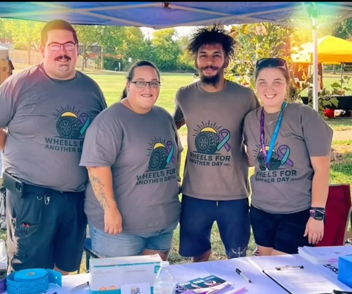 Four people standing together outdoors under a canopy, wearing matching gray t-shirts with a logo for Wheels for Another Day Inc., during a community event.