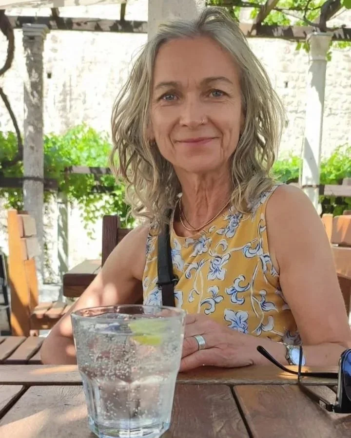 A smiling older woman with gray hair sitting outdoors at a wooden table, with a glass of sparkling water with lime in front of her.