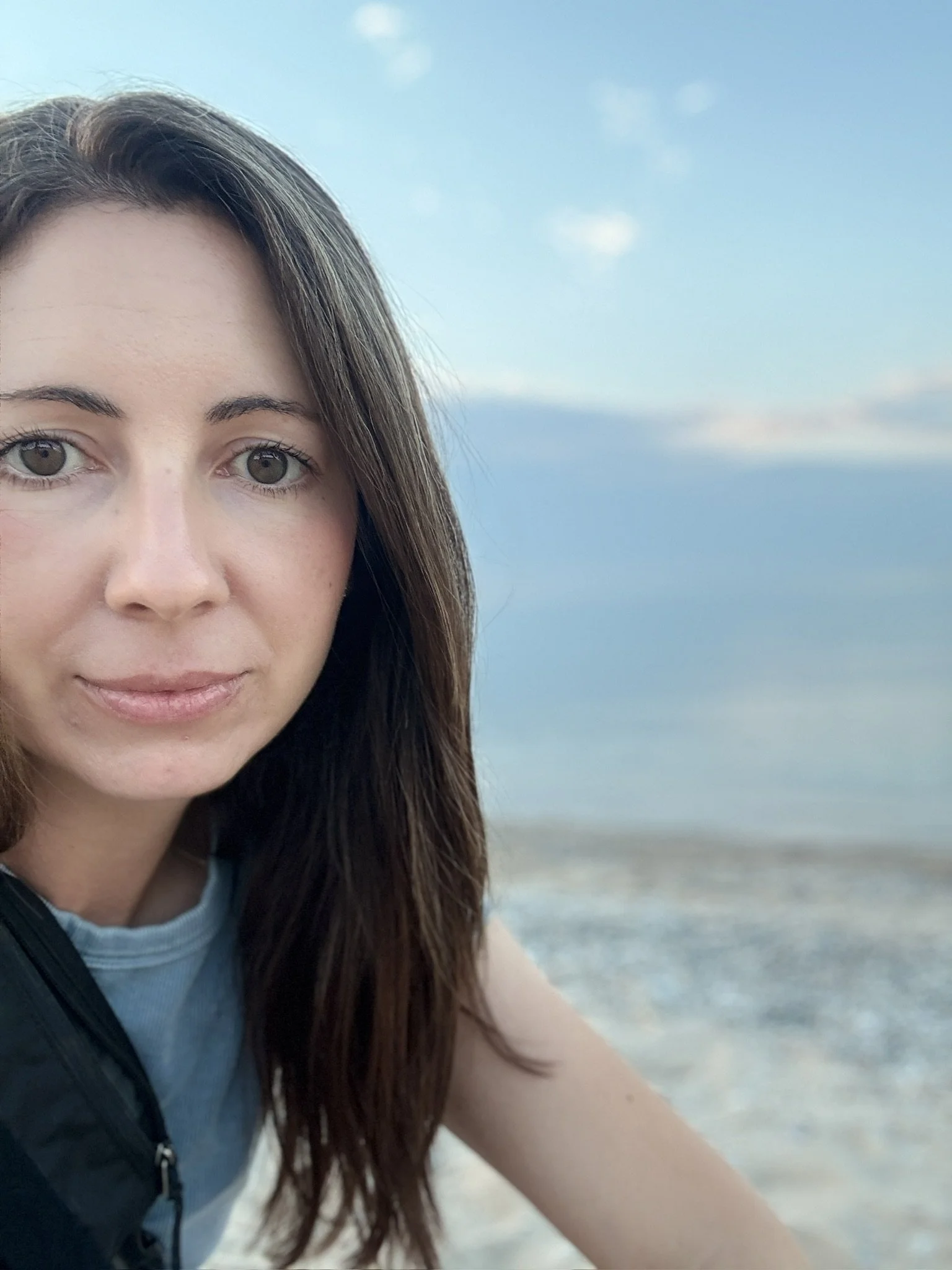 A close-up of a young woman with long brown hair and light-colored eyes at the beach, with the ocean and sky in the background.