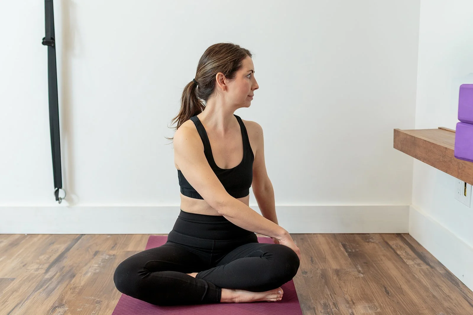 A woman practicing yoga in a seated twist pose on a black yoga mat in a minimalist room with wooden floors and white walls.