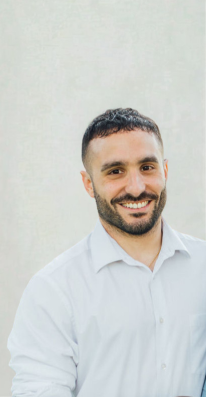 A man with short dark hair and a beard, smiling, wearing a white collared shirt, standing against a plain light-colored background.