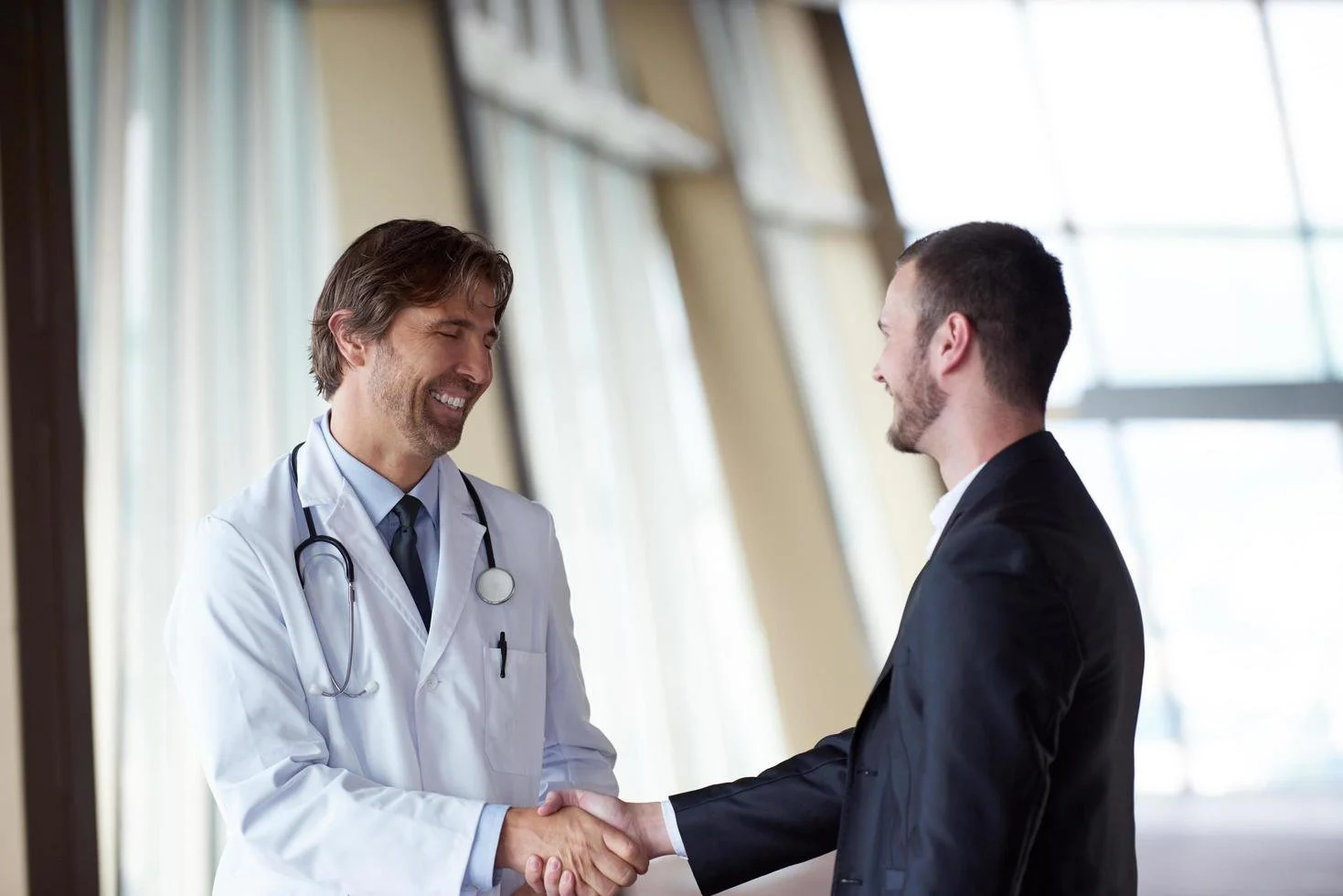A doctor in a white lab coat shaking hands with a man in a black suit, both smiling, indoors with large windows.