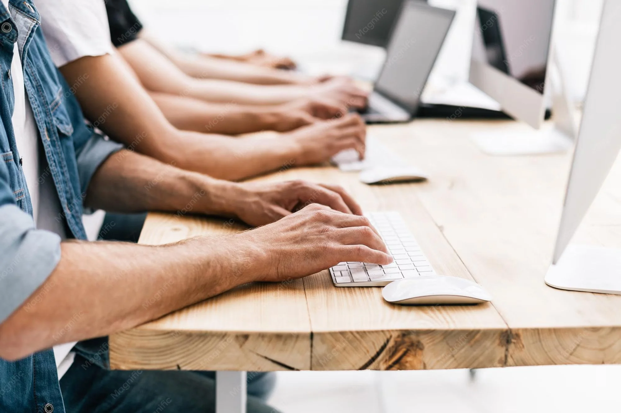 Multiple people working on desktop computers in a row at a shared wooden desk.