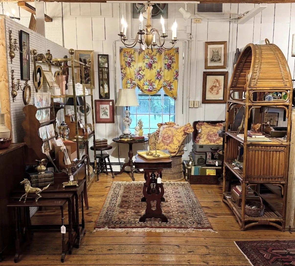 An antique store display with wooden furniture, shelves filled with decorative items, framed artwork, a window with floral curtain, and a chandelier hanging from the ceiling.