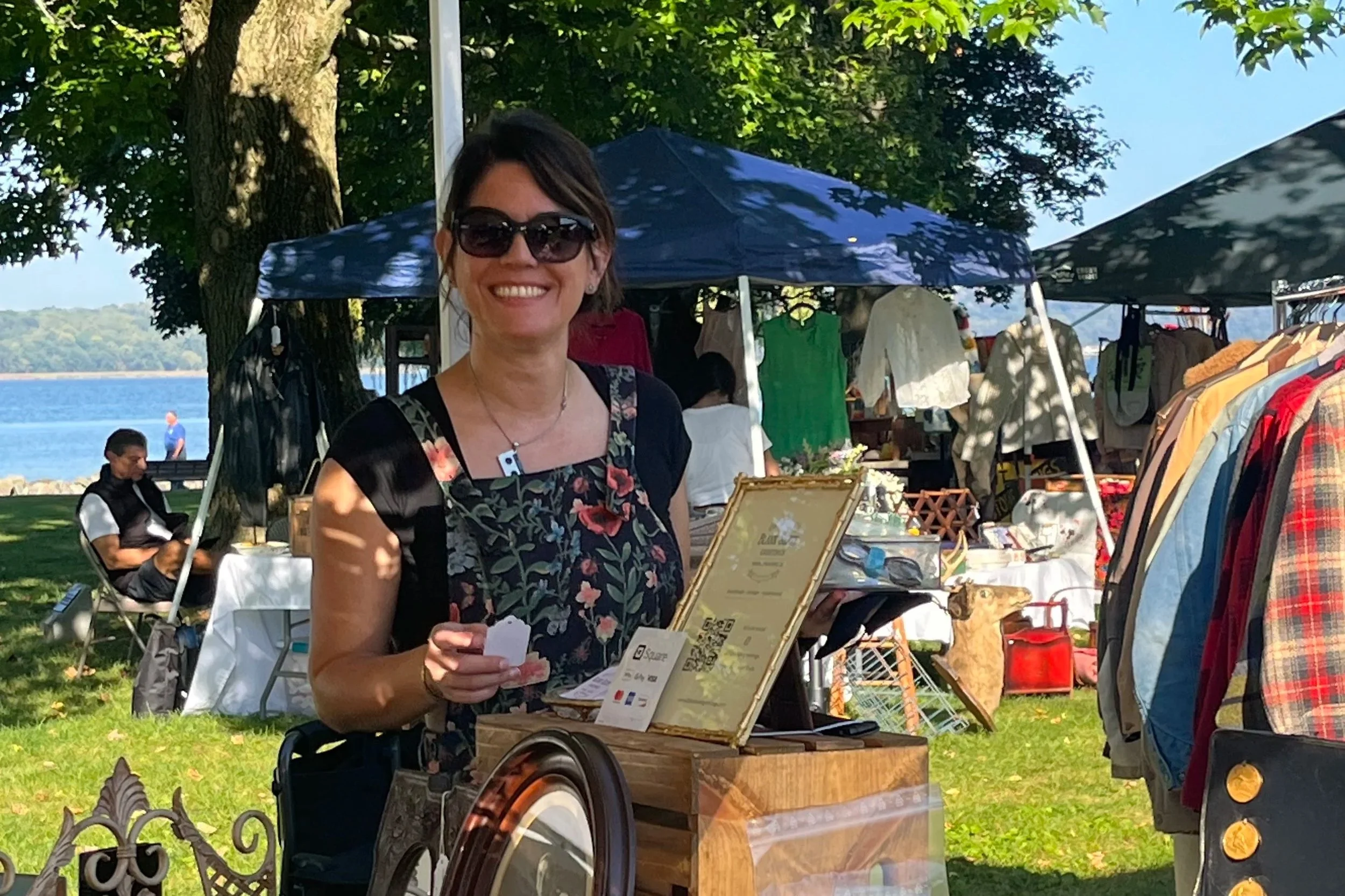 Smiling woman wearing sunglasses and a floral apron at an outdoor market by the Hudson River on a sunny day.