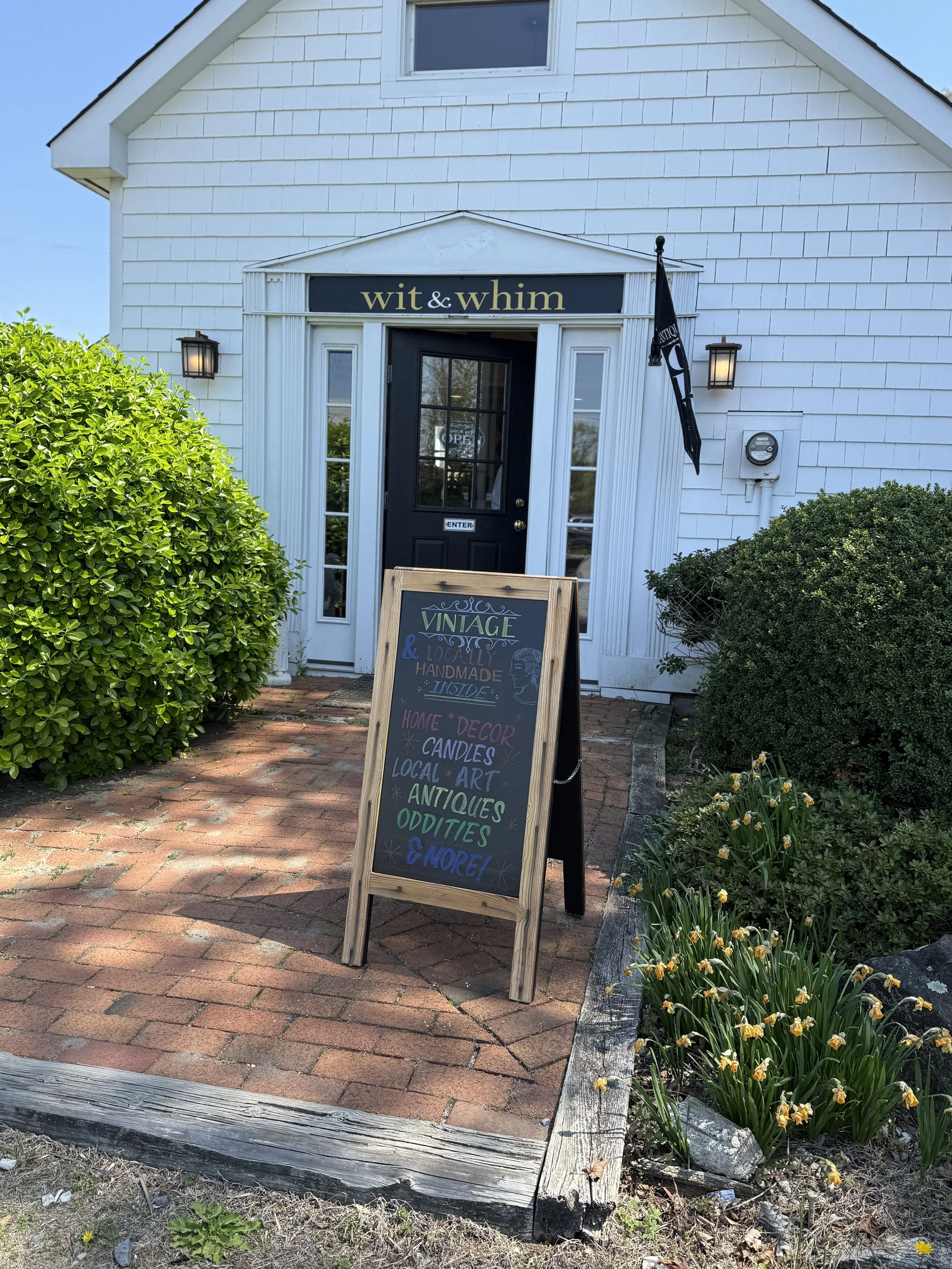 Exterior of a small white storefront with a black door and a chalkboard sign in front, surrounded by green bushes and yellow flowers.