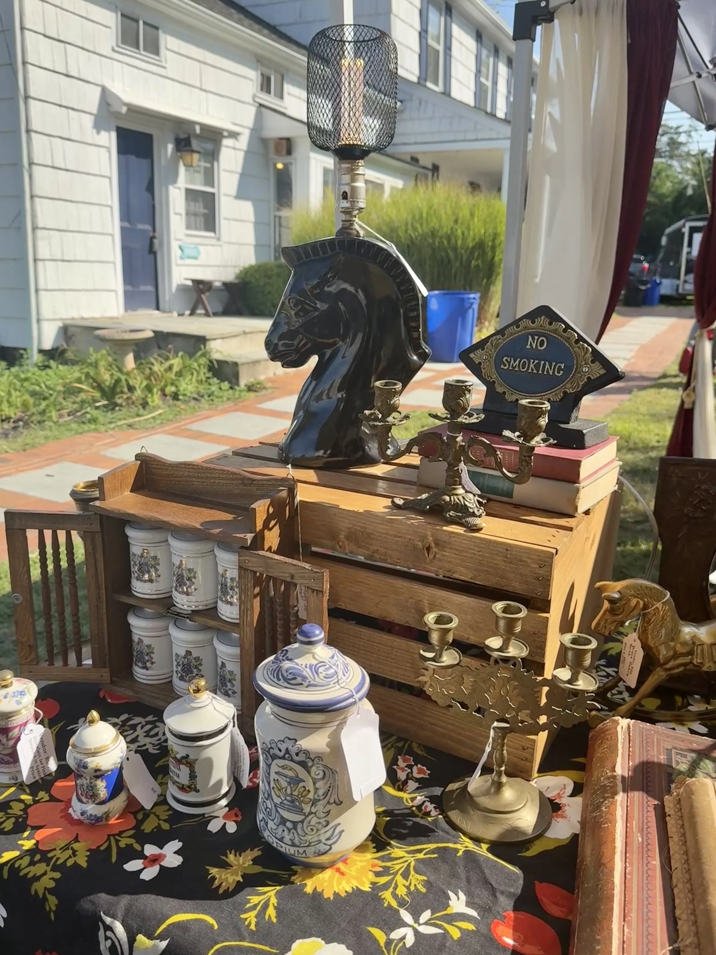 Outdoor flea market display with vintage items including ceramic jars, a black horse head lamp, brass candlesticks, a horse figurine, and a "No Smoking" sign, set against a residential background.