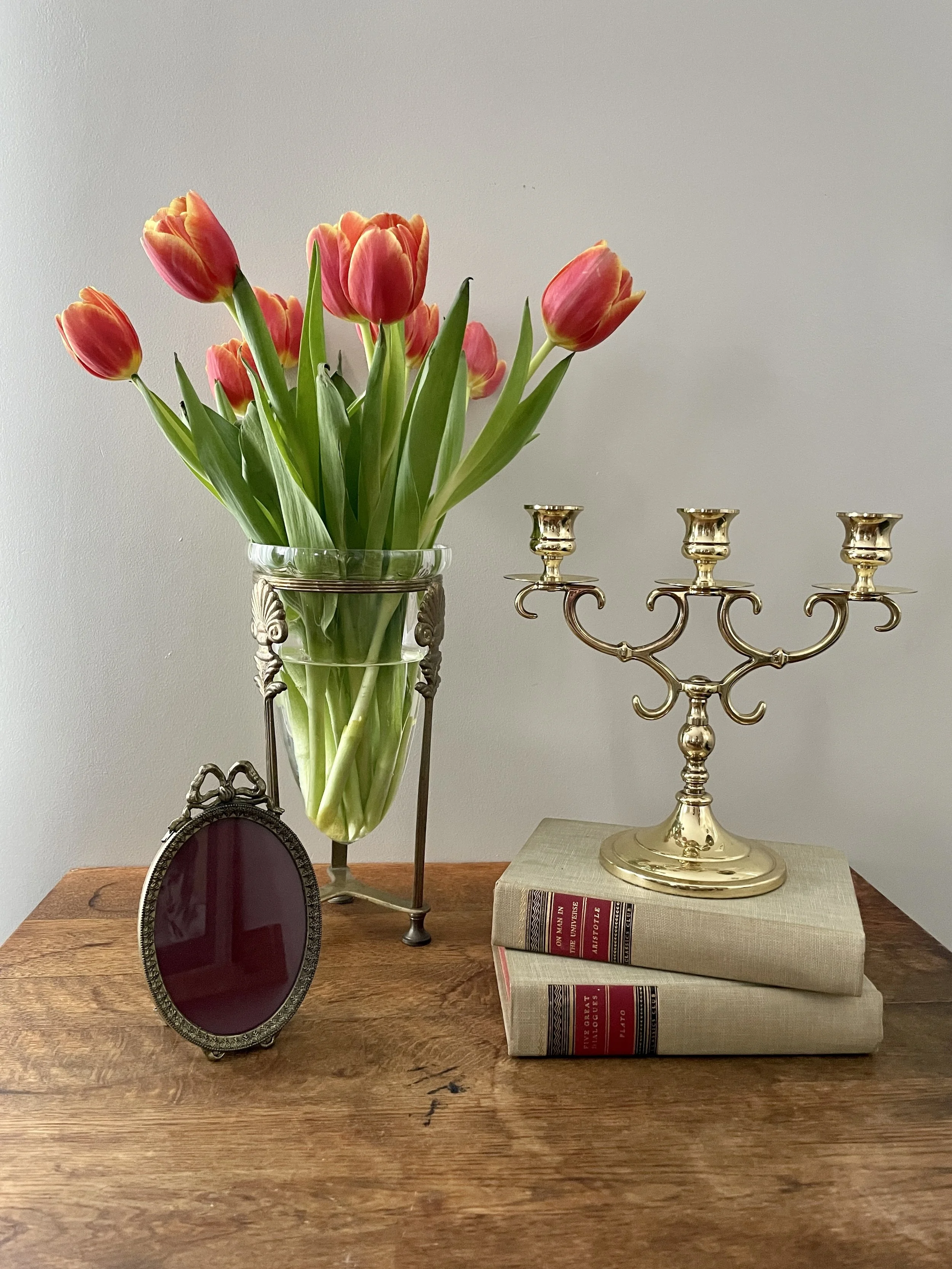 A wooden table with a glass vase of pink and yellow tulips, a gold-colored candelabra on top of stacked books, and an oval picture frame with a red background.
