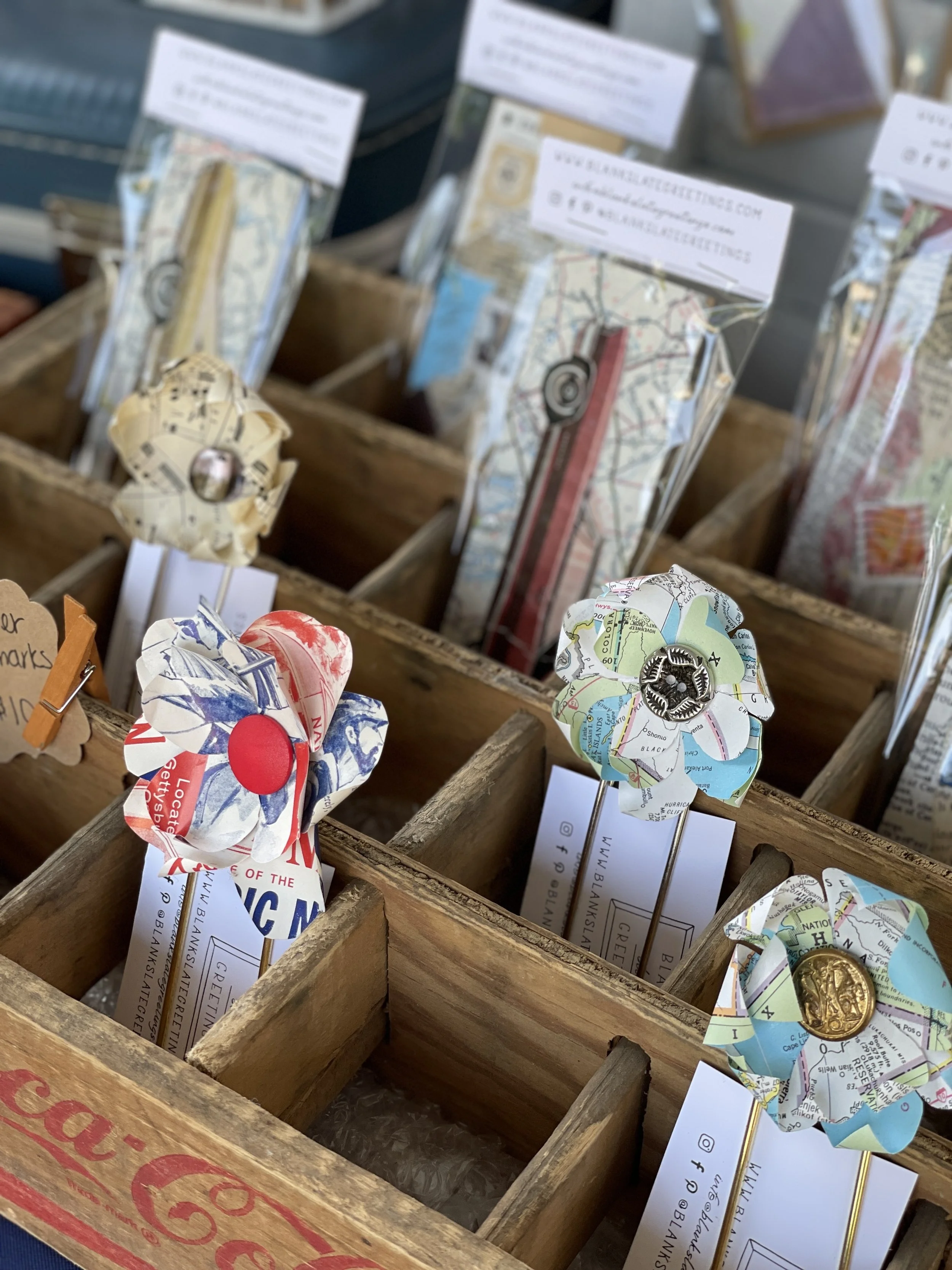Colorful paper flower pins with buttons and map patterns in a wooden display box.