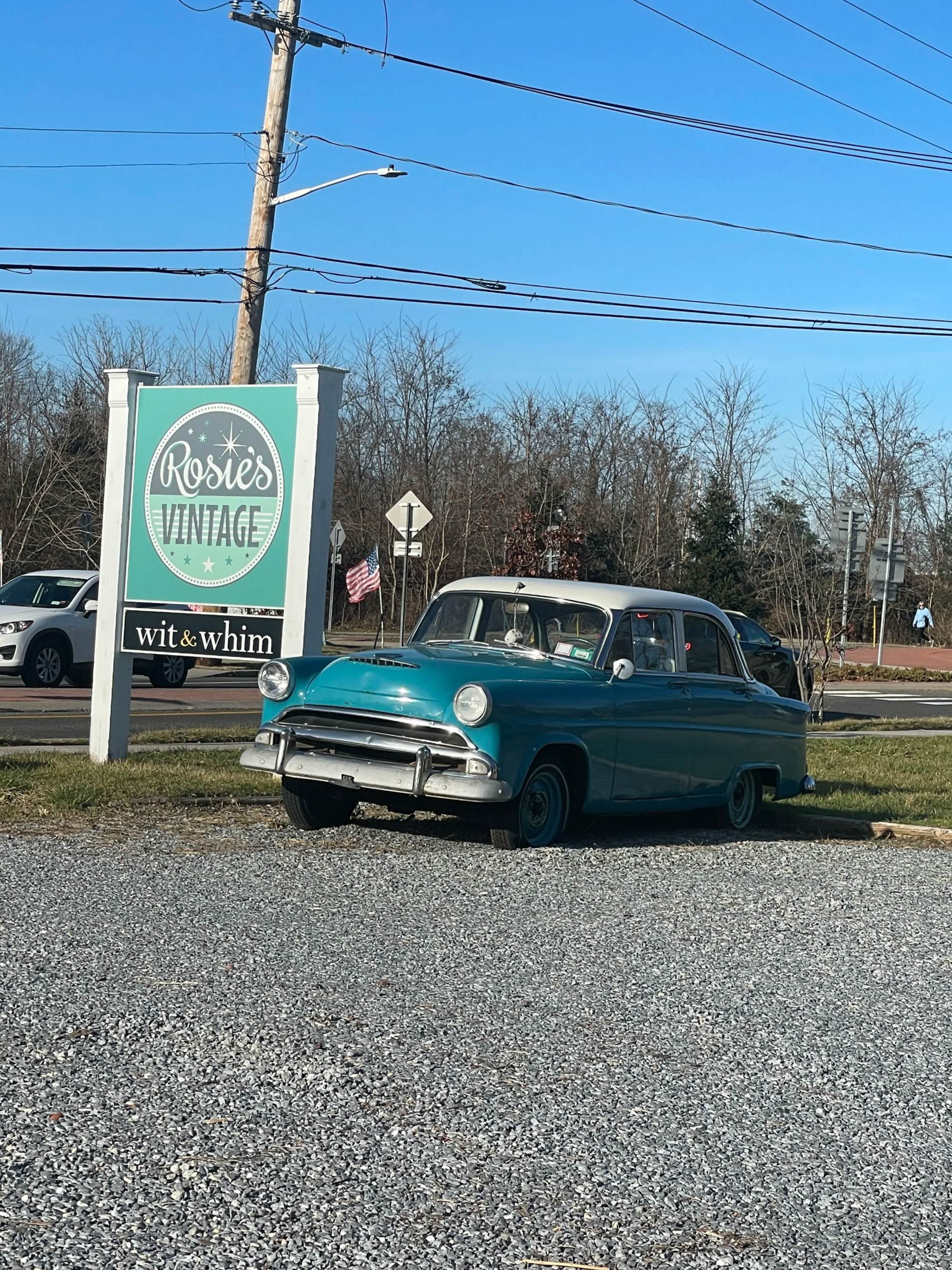 Vintage blue car parked in front of a sign for Rosie's Vintage in a parking lot with gravel, under power lines, with trees and a clear blue sky in the background.