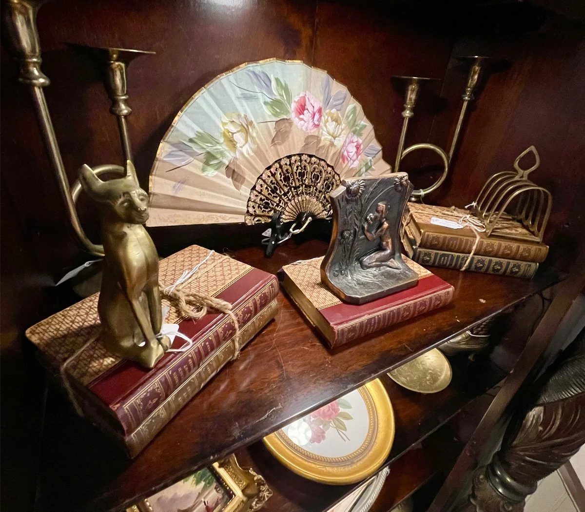 Decorative objects on a wooden shelf, including a brass cat figurine, an ornate handheld fan with floral design, a small sculpture of a woman, vintage books, a brass letter opener, a decorative plate with flowers, and a framed picture.