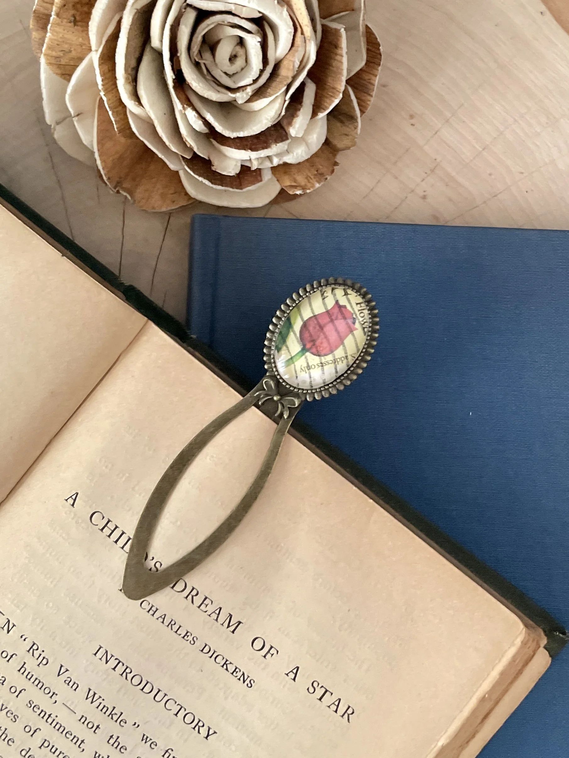 An open book titled "A Child's Dream of a Star" by Charles Dickens, a blue notebook, a decorative bookmark with a rose postage stamp illustration, and a wooden flower sculpture on a wooden surface.