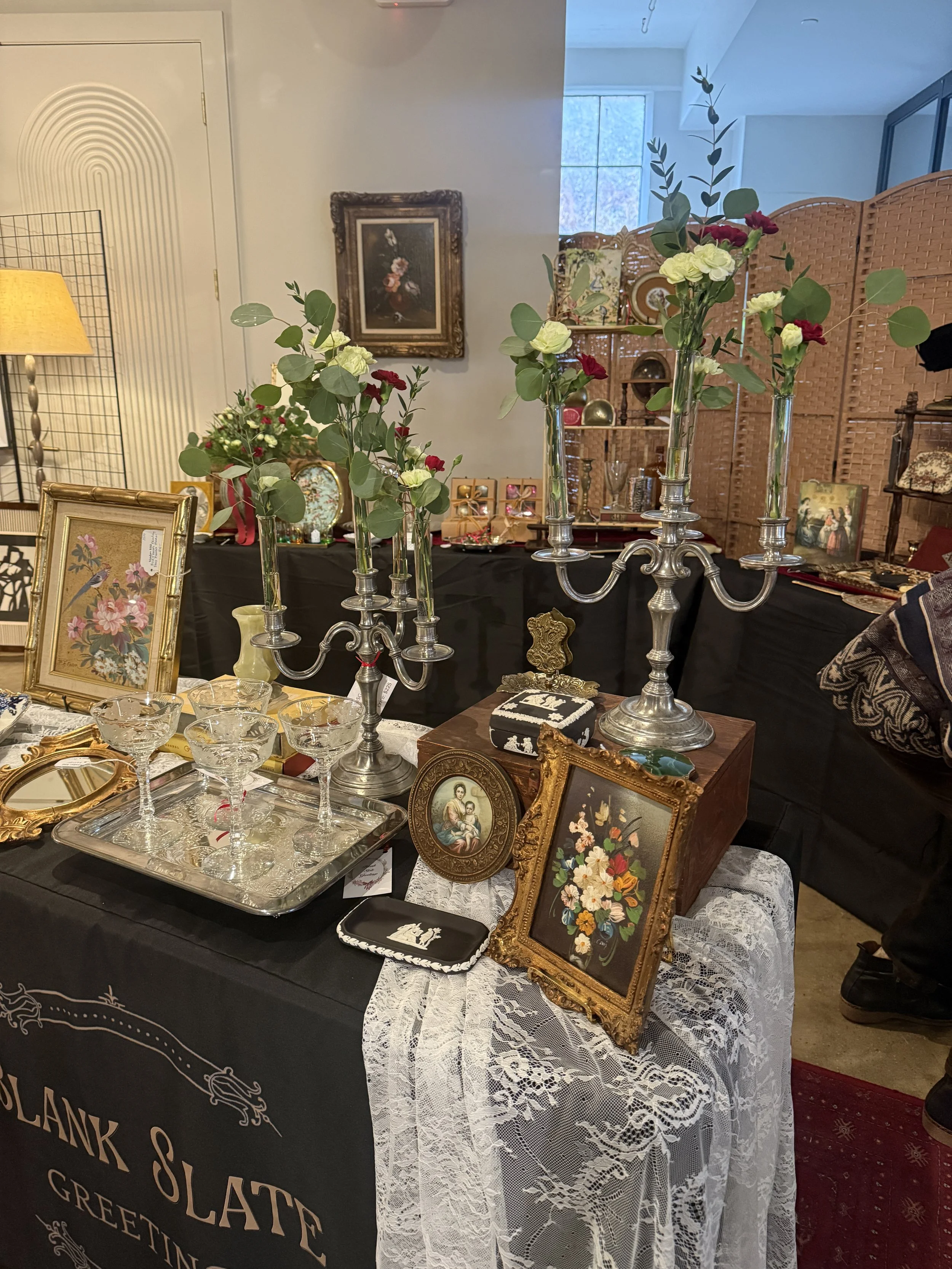 A table displays vintage picture frames, glassware, floral arrangements in tall vases, decorative items, and assorted small objects at an antique sale or display, set against a backdrop of a room with artwork and shelving.