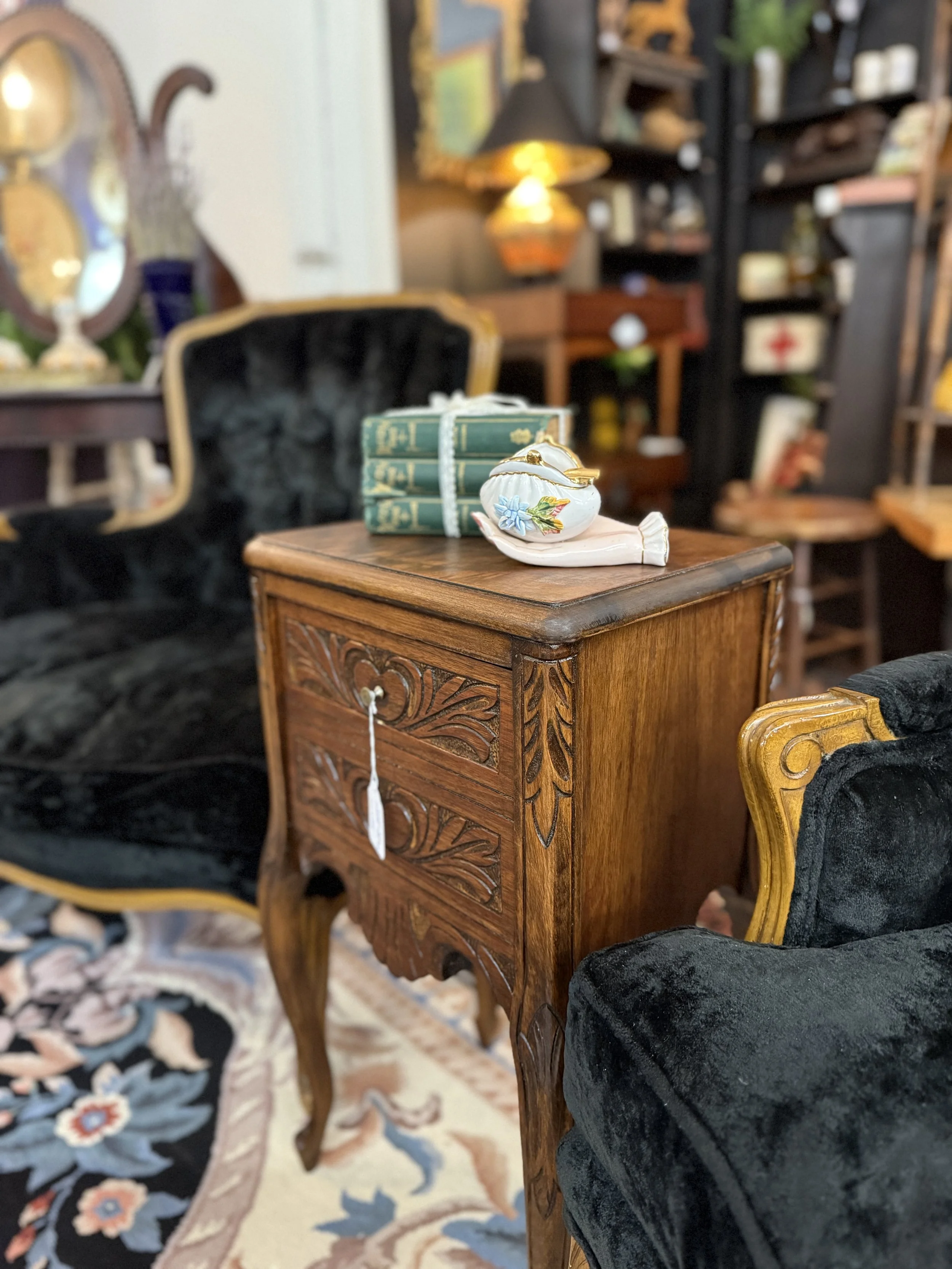 An antique wooden side table with carved details, placed between two black velvet armchairs with gold trim, in a vintage furniture store or room.