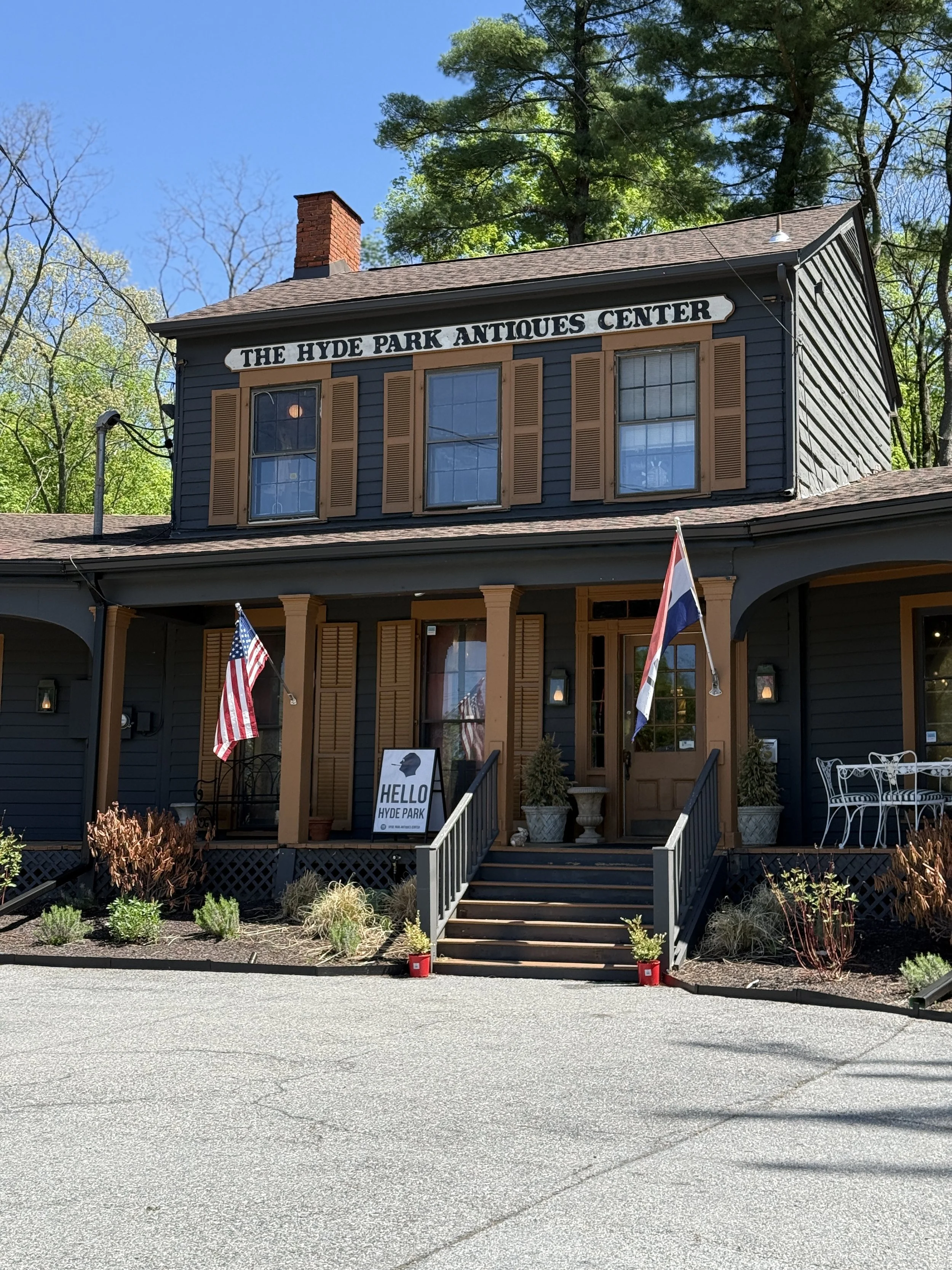 Front view of The Hyde Park Antiques Center, a black two-story building with brown shutters and a porch, decorated with American and Dutch flags, surrounded by trees and shrubs.
