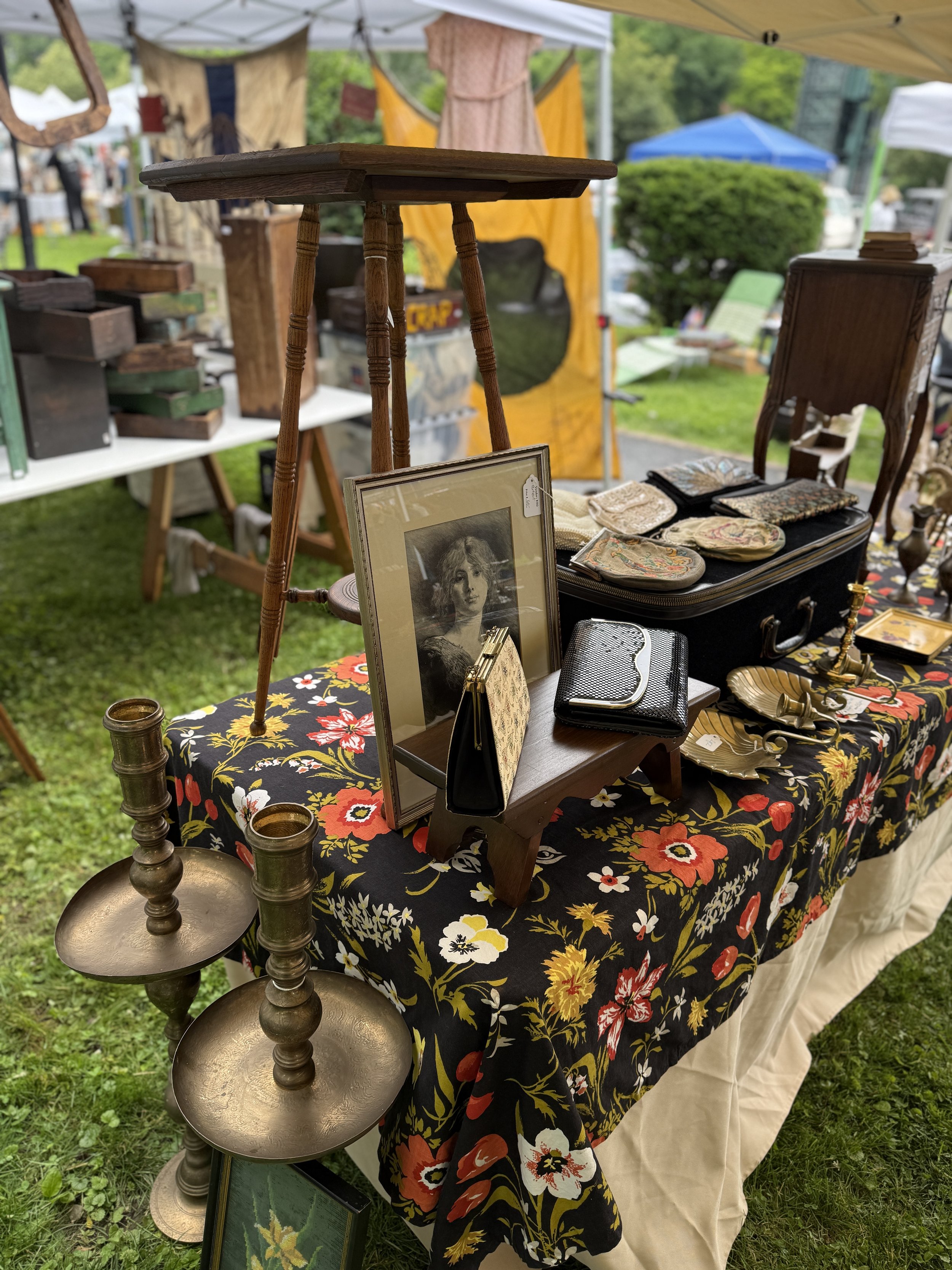 A flea market stall displays vintage items including a framed portrait of a woman, small handbags, a black suitcase with fabric pouches, candle holders, and decorative trinkets on a floral tablecloth.