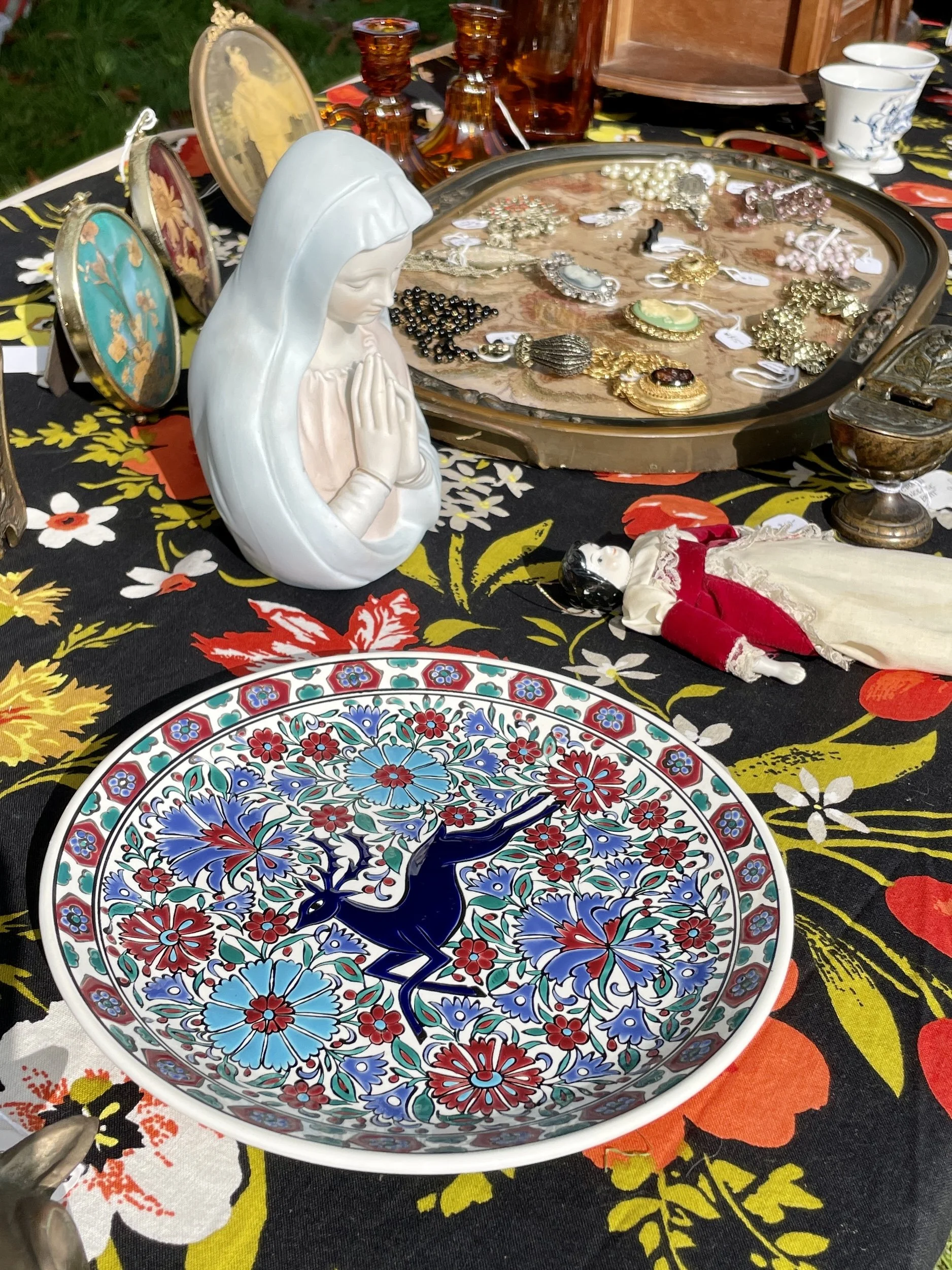A table displaying various vintage items, including a decorative plate with a blue deer motif, a praying Madonna figurine, a doll in a dress, several jewelry pieces, and glassware, all arranged on a floral tablecloth.