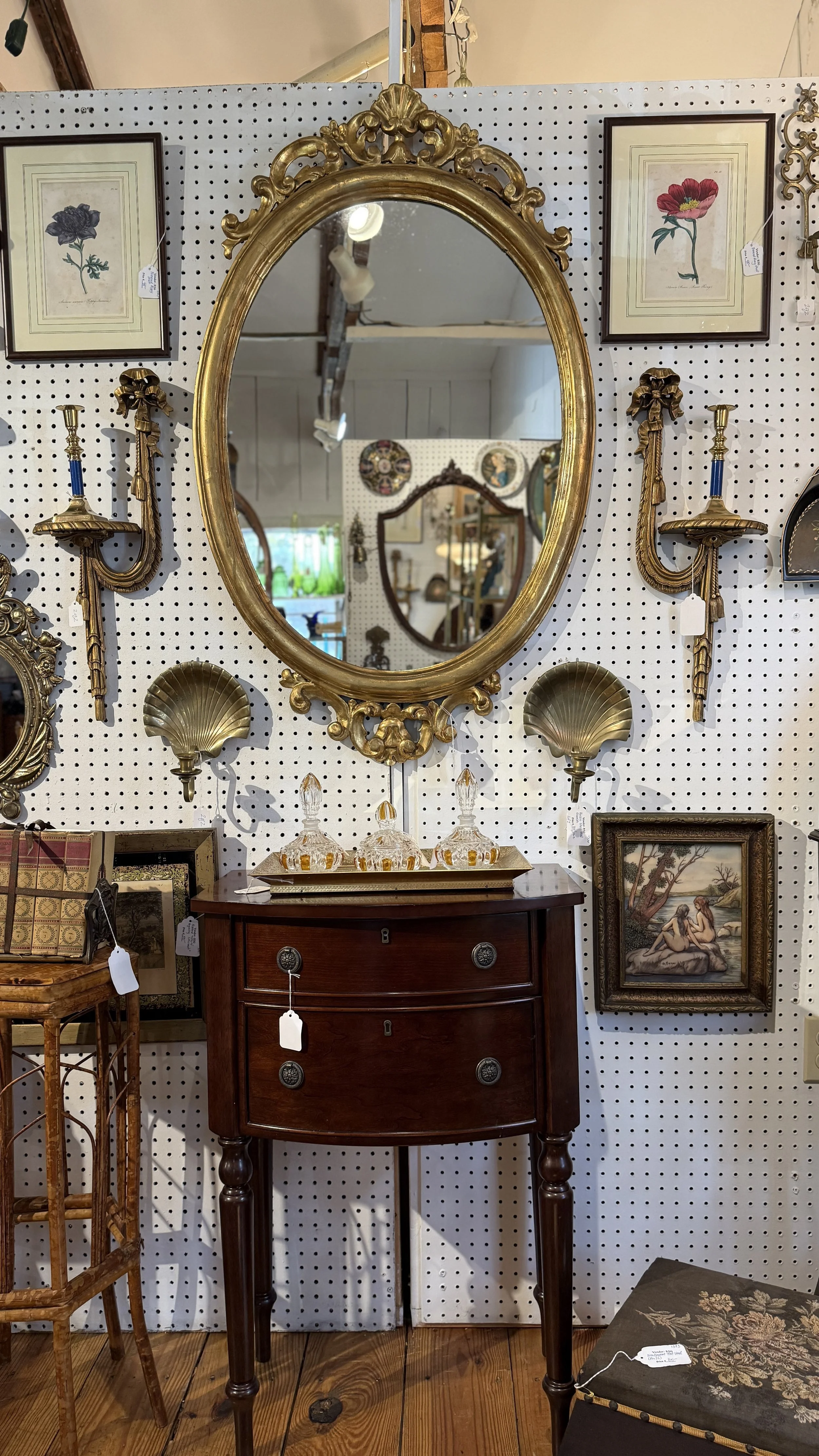 An antique wooden dresser with a curved front, four legs, and decorative silver handles, topped with a gold-colored tray holding glass decanters. Above it is a large ornate gold-framed oval mirror, flanked by two wall-mounted candle sconces and frame