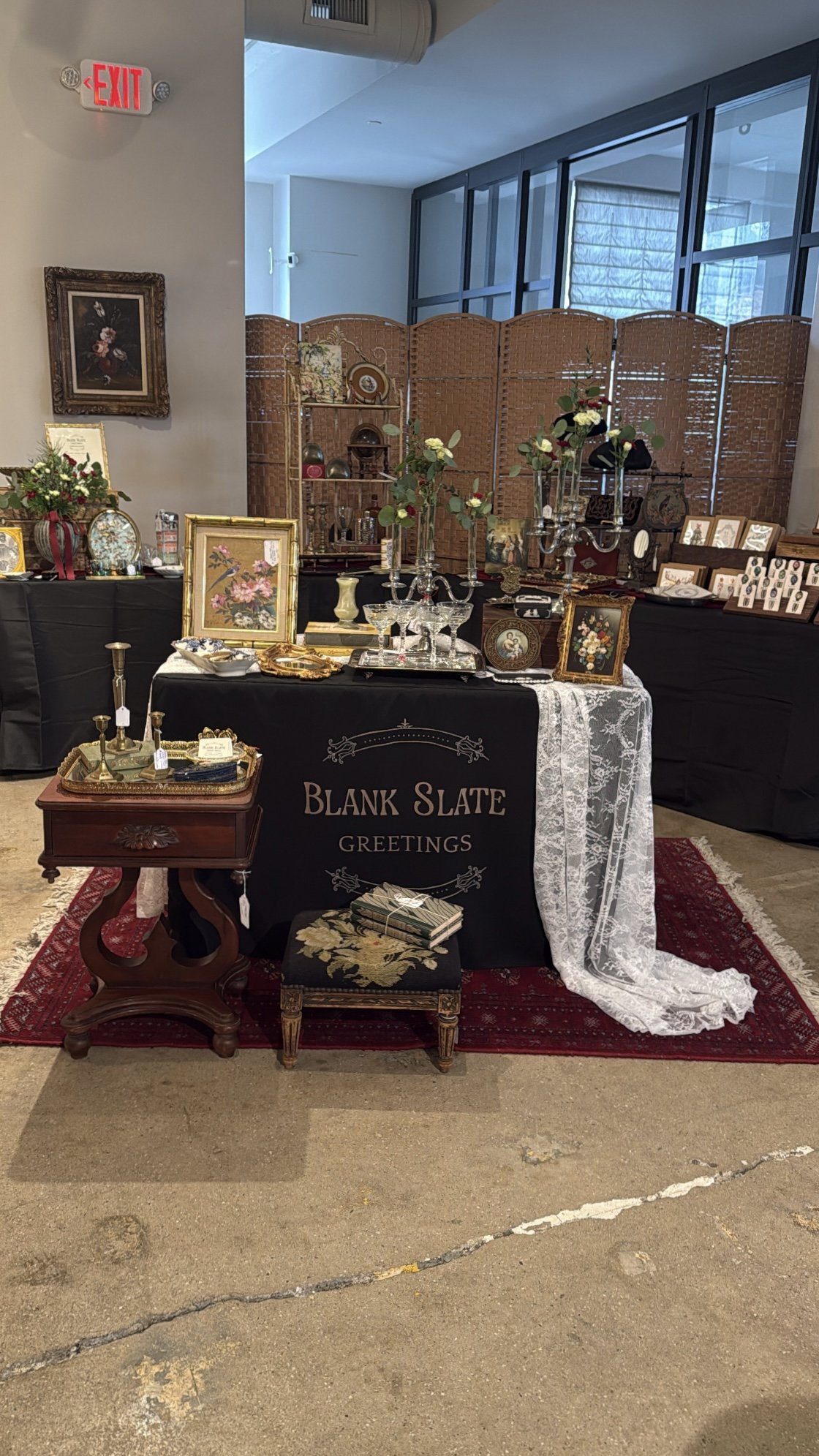 An antique and vintage items display at an indoor market stall with a black tablecloth that reads 'Blank Slate Greetings.' Items include framed artwork, silver candelabras, glassware, jewelry, and decorative trinkets on and behind the table.