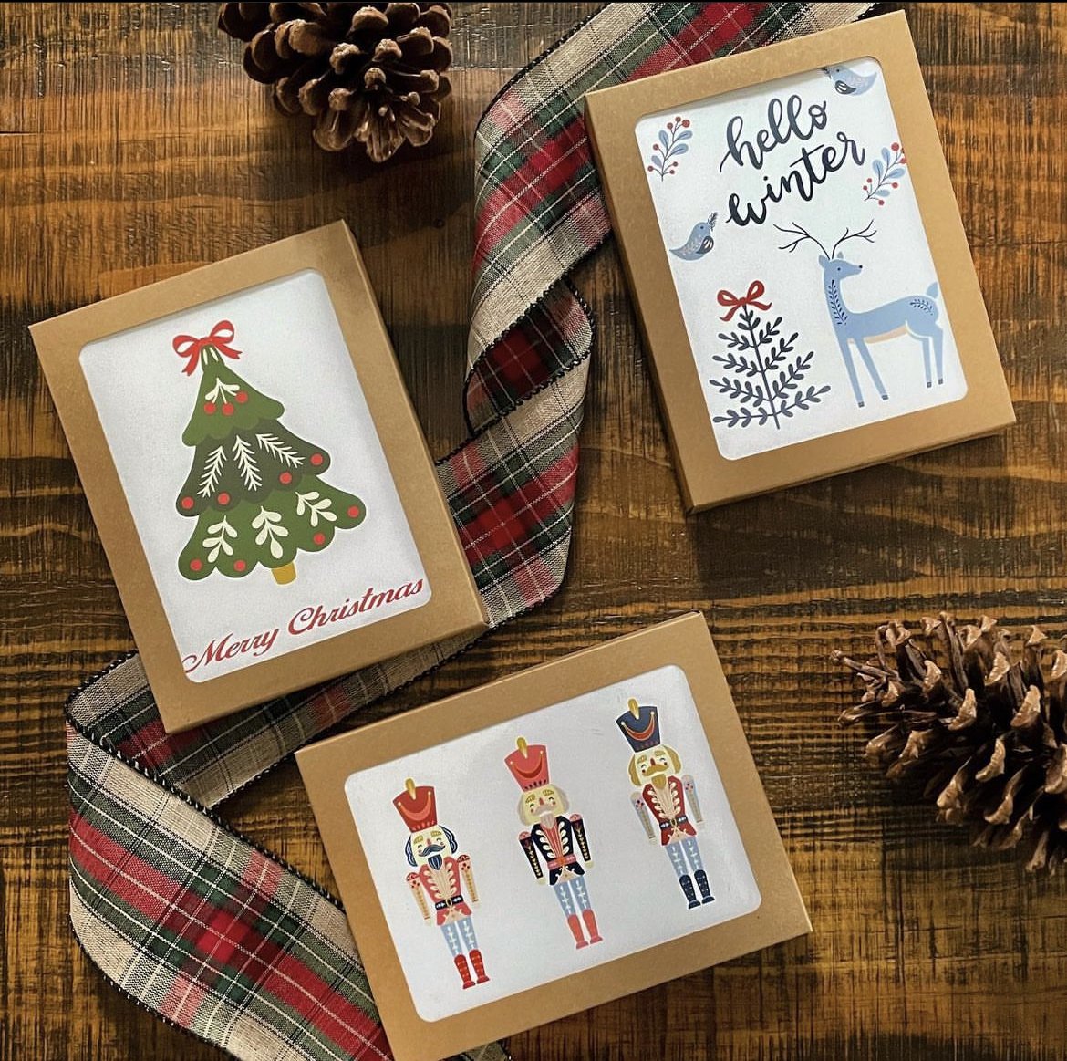 Three Christmas-themed gift boxes on a wooden table, decorated with holiday illustrations and ribbons. One box features a Christmas tree with the words "Merry Christmas," another has a winter scene with a reindeer and birds and the words "hello winte