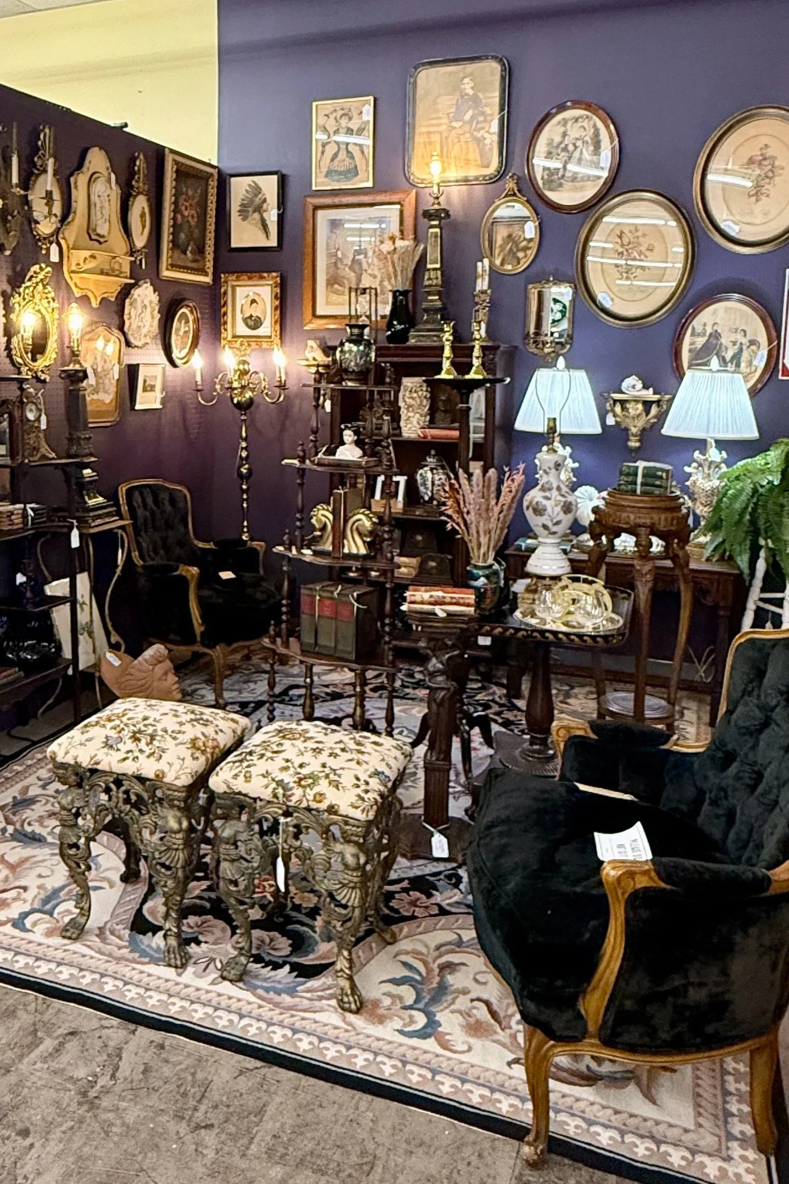 An antique furniture and decor display with vintage chairs, framed artwork on purple walls, ornate table lamps, and decorative accessories in a vintage store.