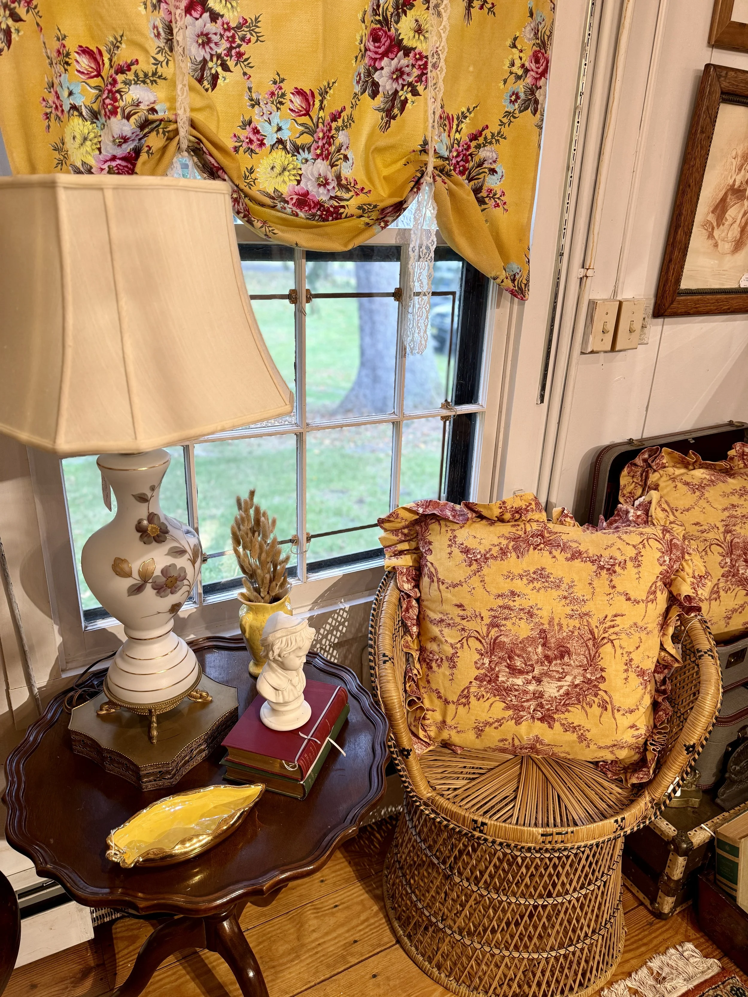 A cozy corner with a window curtain featuring a floral pattern in yellow, pink, and purple. A wicker chair with yellow and pink floral cushions is next to a wooden side table holding a white flower vase, a small white bust, a yellow dish, two books, 