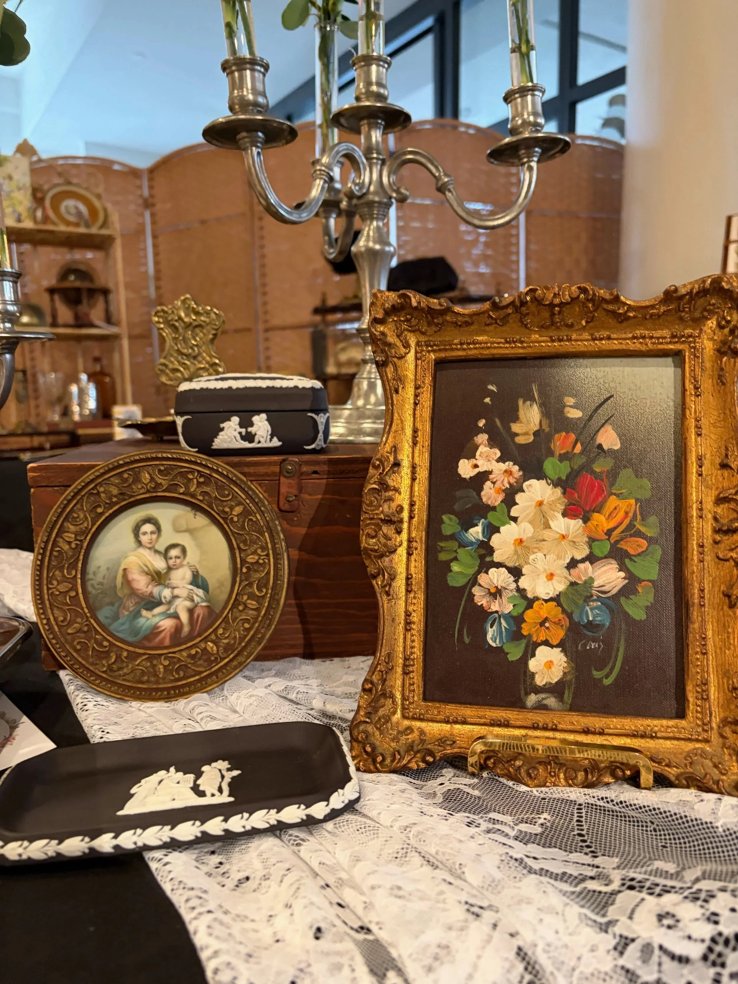 Various vintage decorative items including framed artwork, a black and white brooch, and a silver candelabra displayed on a lace tablecloth at an antique store.