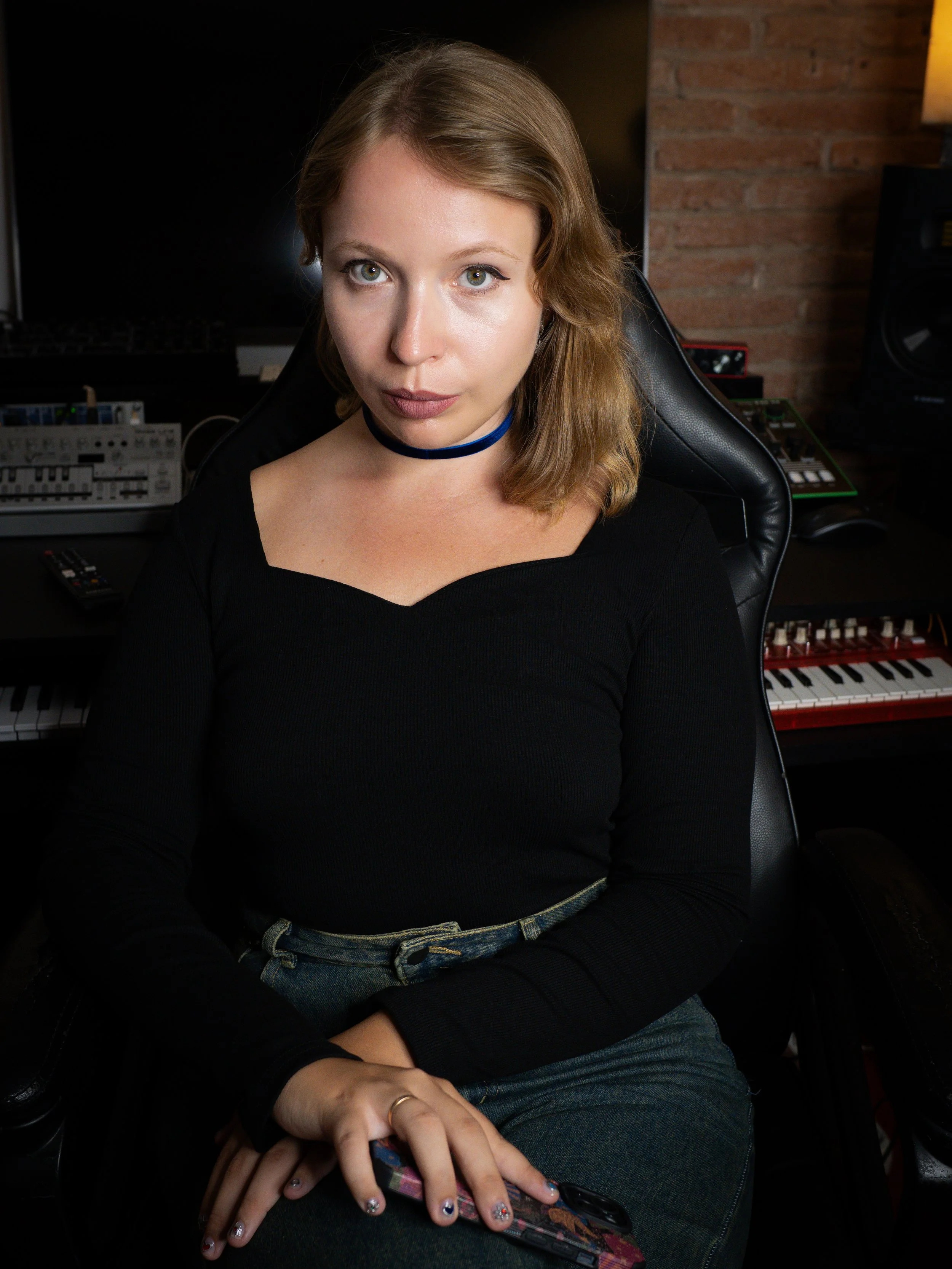 A young woman with shoulder-length red hair and blue eyes sitting in a black chair in a music studio, holding a smartphone with a colorful case, with music equipment and a brick wall in the background.