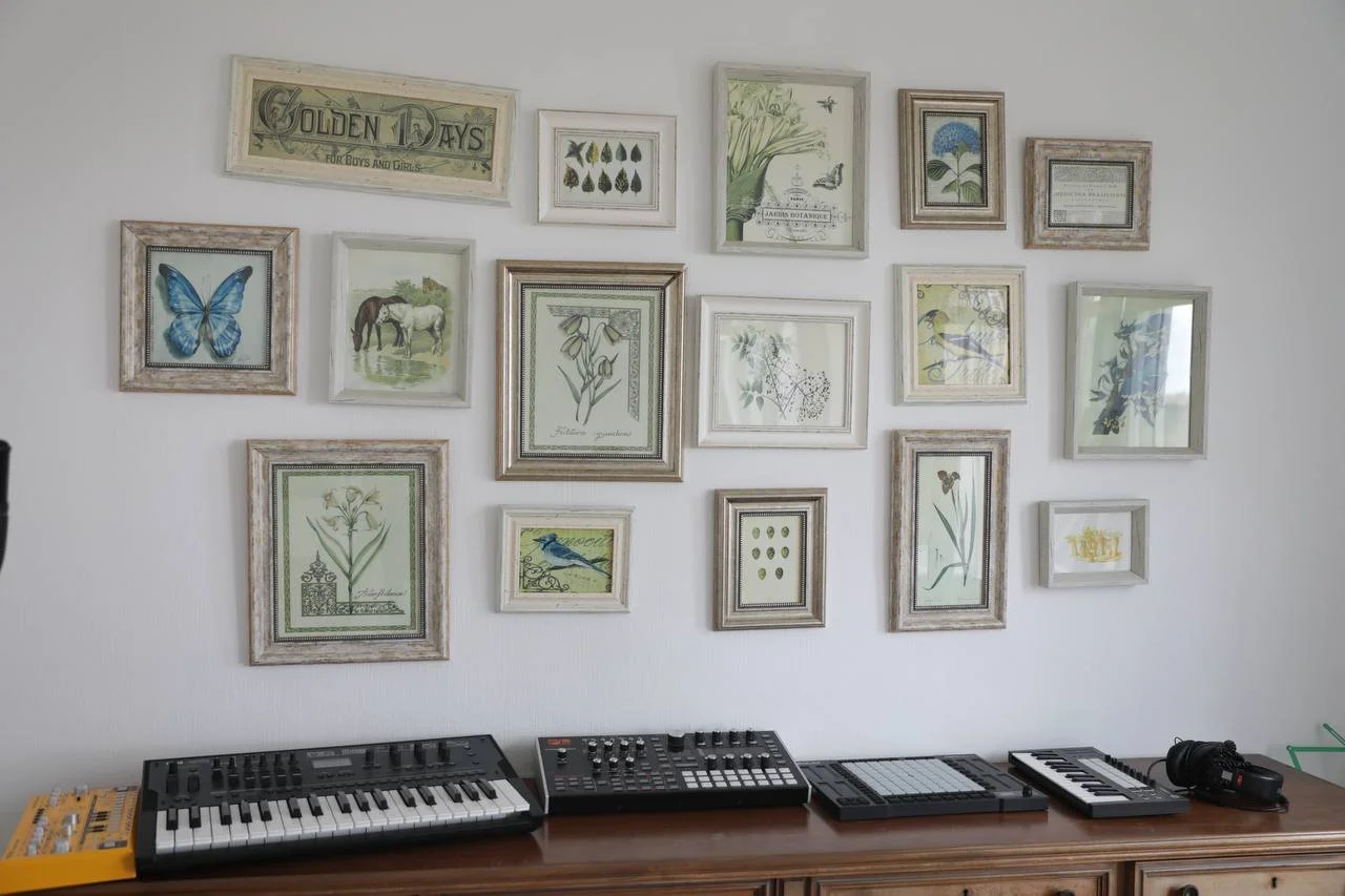 Music production setup with three electronic keyboards, a mixer, and a pair of headphones on a wooden table, with a white wall decorated with framed botanical and nature-themed artwork.