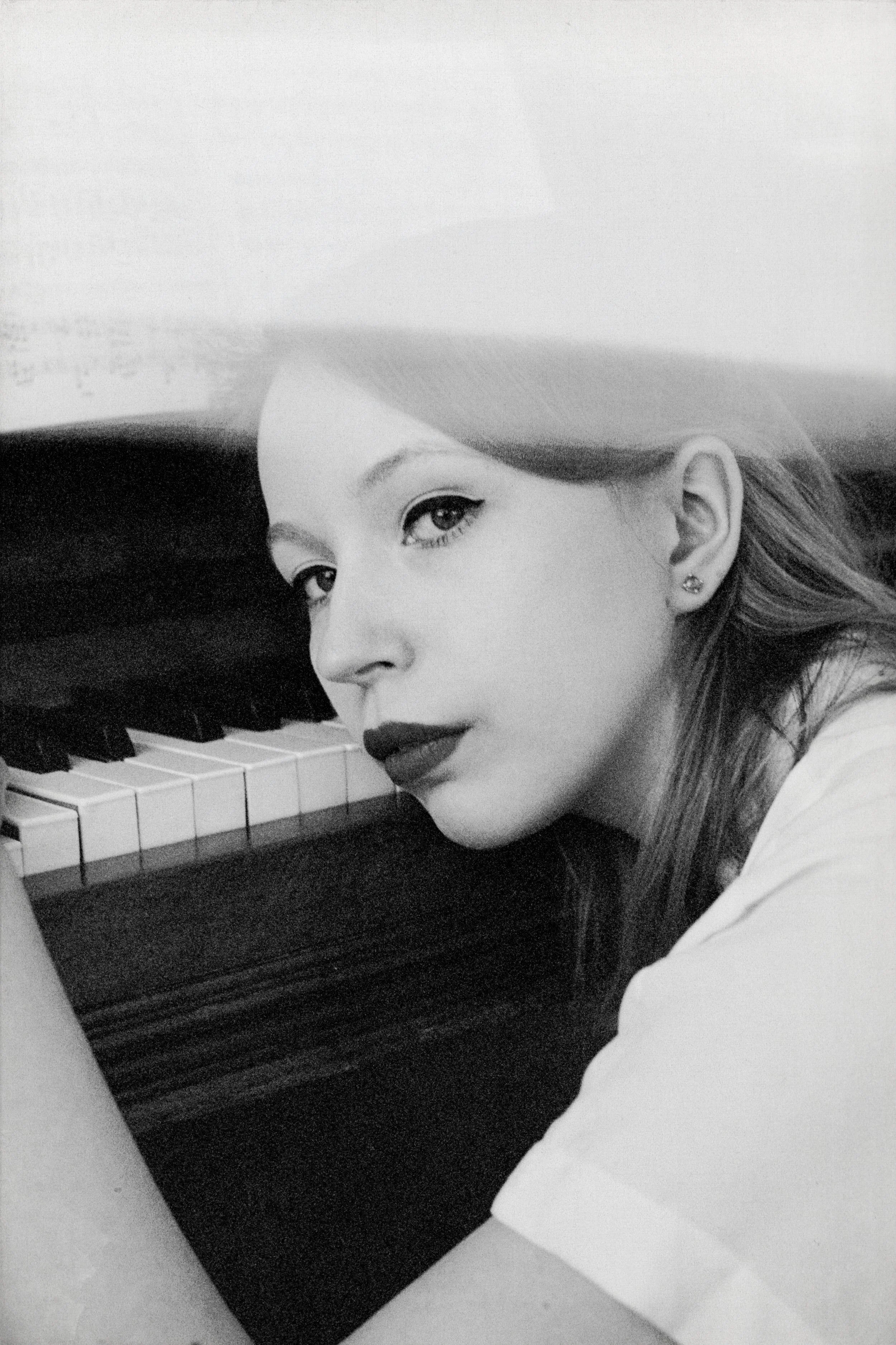 Black-and-white photo of a young woman with long hair, wearing makeup, and earrings, leaning on a piano with her face close to the keys.