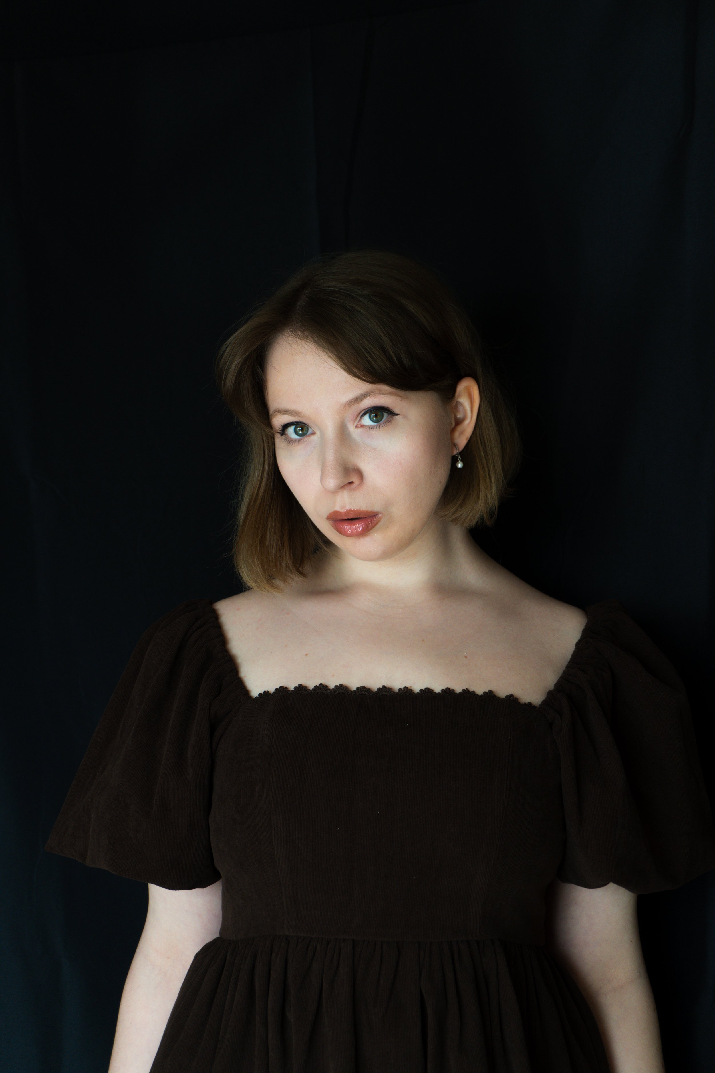 A woman with shoulder-length brown hair and blue eyes wearing a dark brown dress with puffed sleeves and looking at the camera against a dark background.