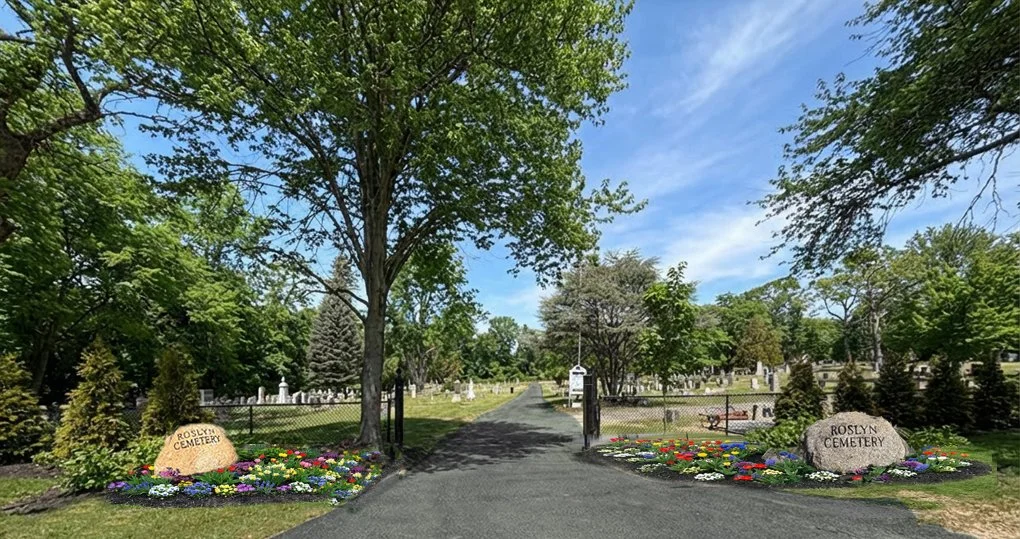 Entrance to Roslyn Cemetery with paved path, trees, colorful flower beds, and tombstones in the background, under a partly cloudy sky.