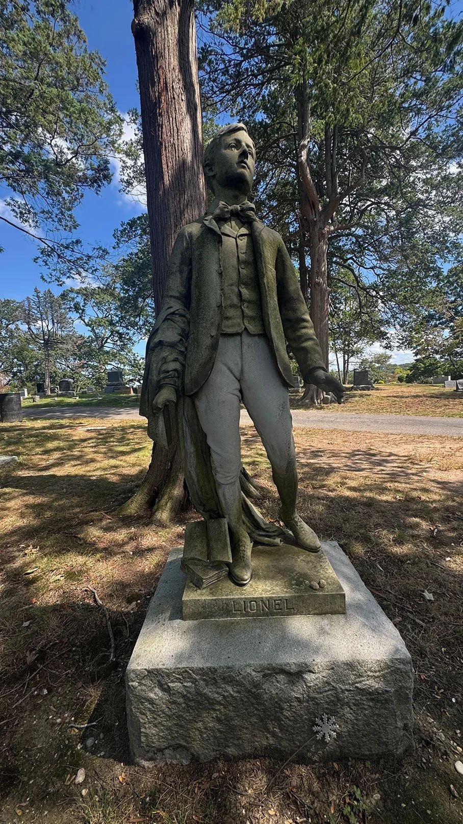 Statue of a young boy standing outdoors in front of a large tree, with a sign reading 'LIONEL' at the base.