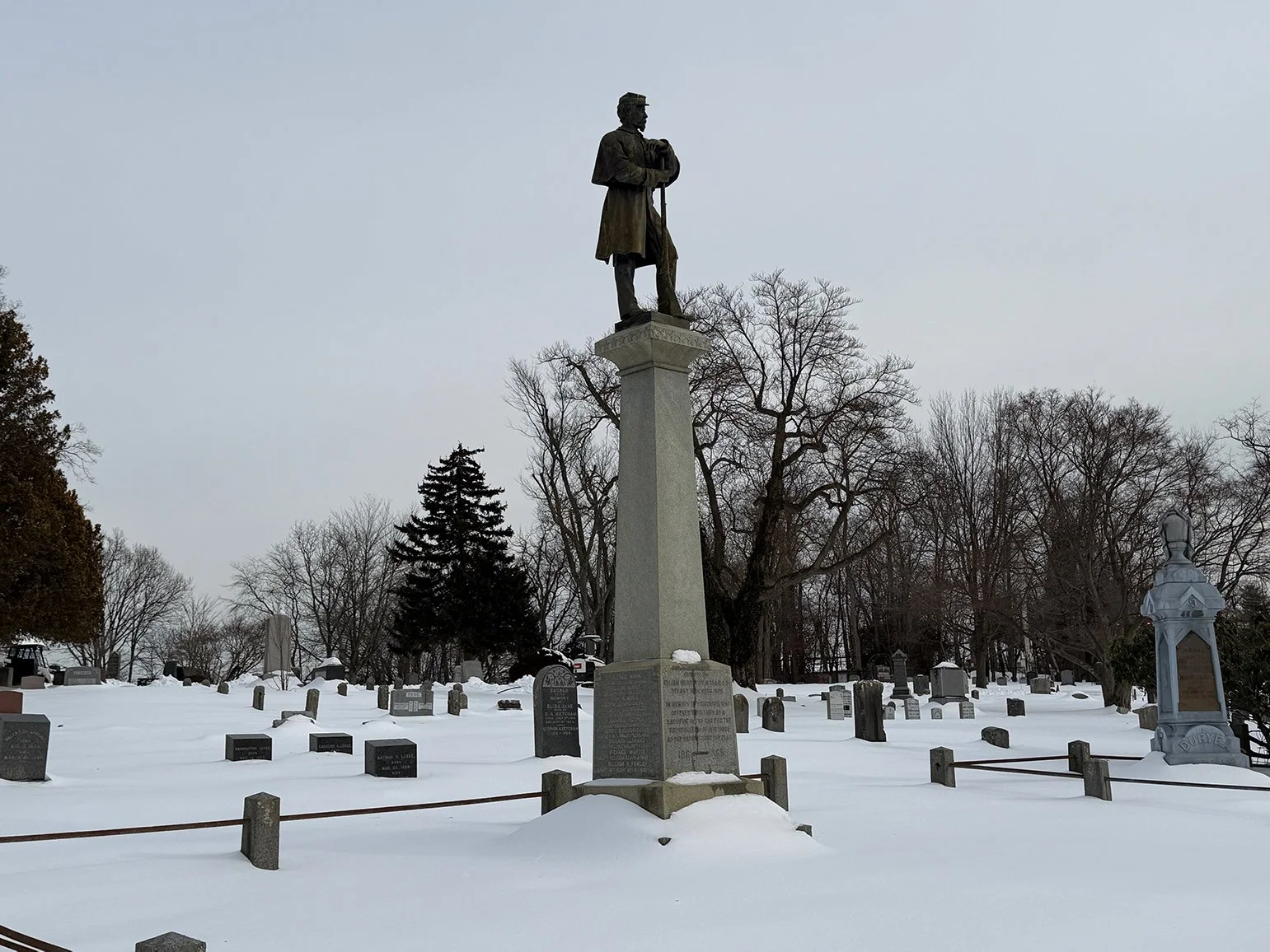 A cemetery with numerous gravestones and monument markers, covered in snow, with a large statue of a historical figure on a tall pedestal in the center, trees without leaves in the background, and an overcast sky.
