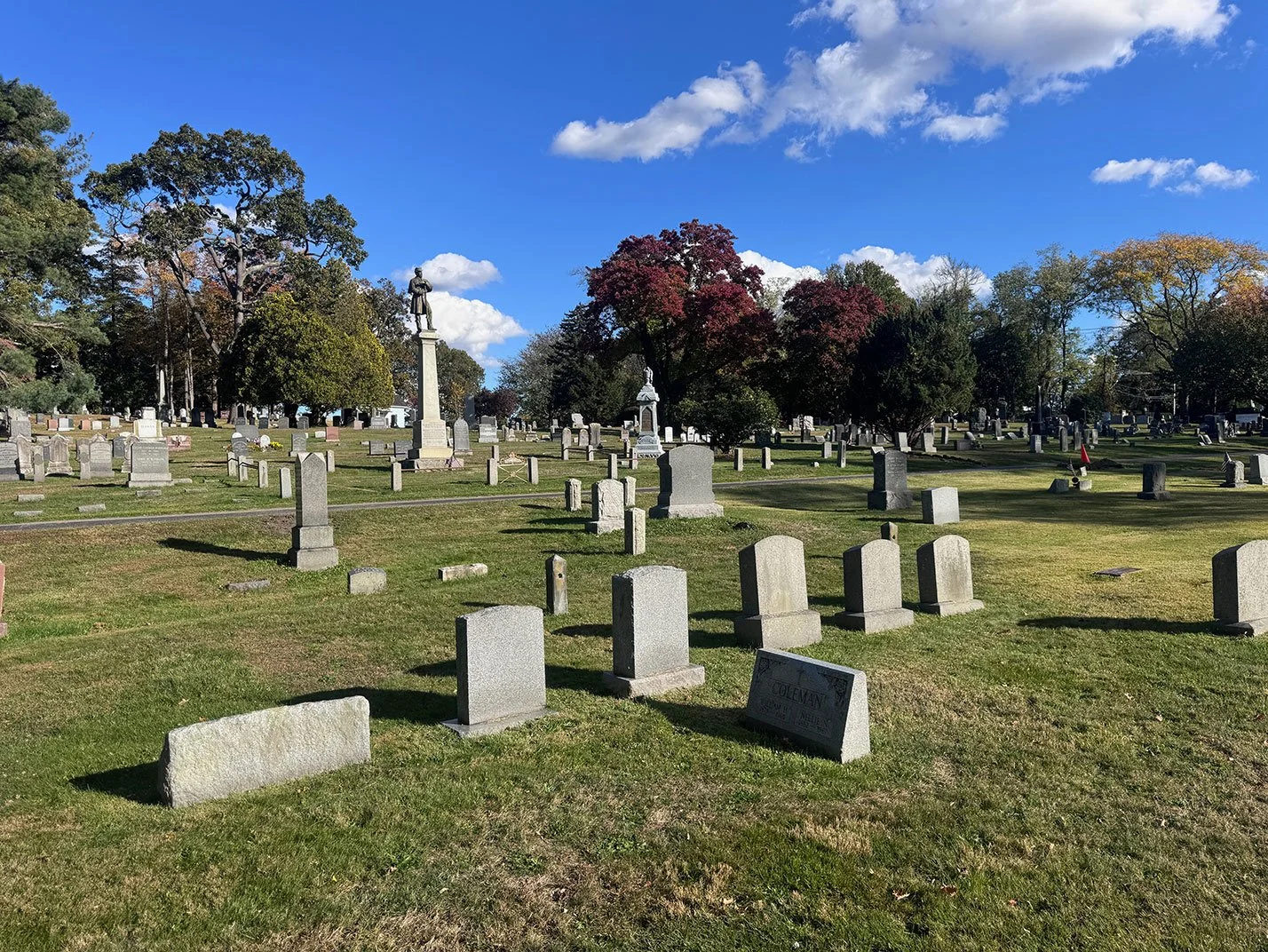A cemetery with numerous headstones and memorials on a bright, sunny day with a clear blue sky and some scattered clouds. There are trees with green and fall-colored leaves surrounding the area.