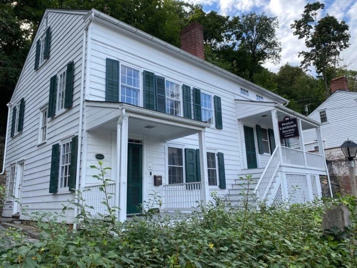 White two-story house with green shutters and front porch, surrounded by greenery, with a sign for the Roslyn Landmark Society.