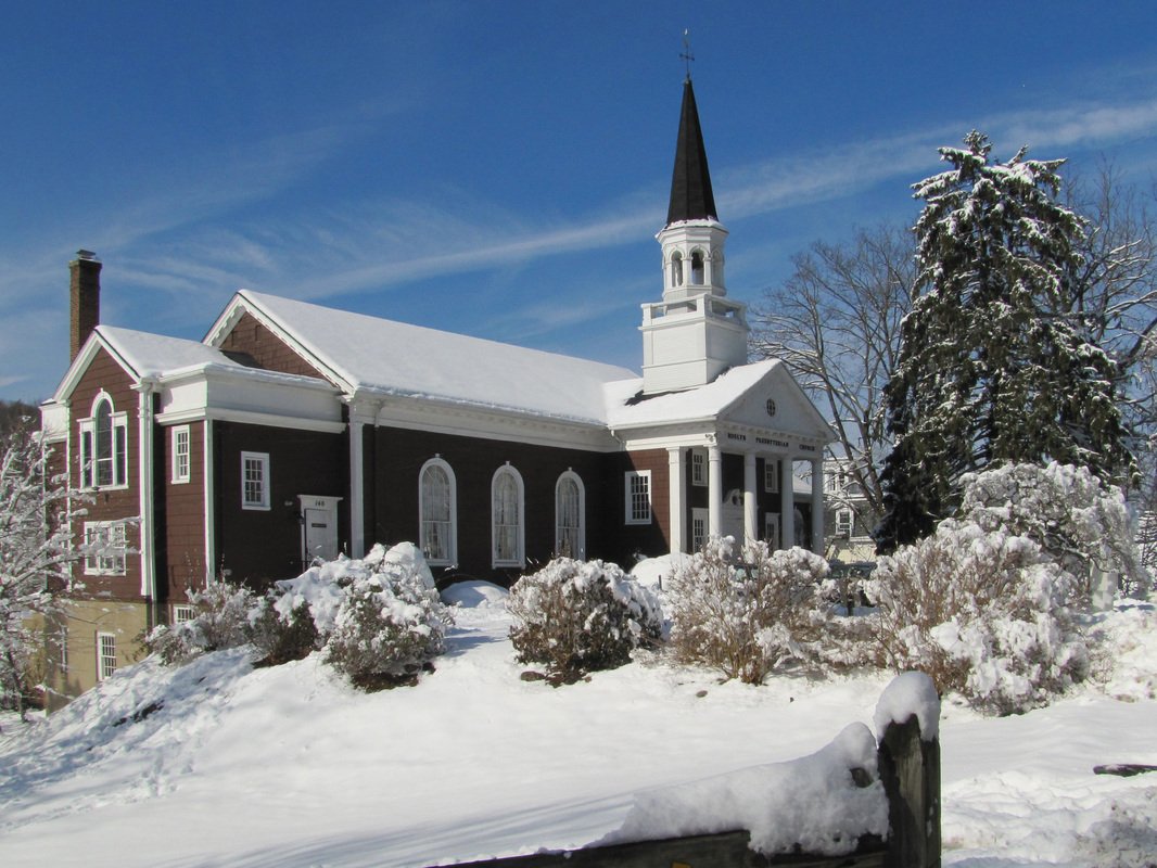 A church building with a steeple surrounded by snow-covered bushes and trees on a bright winter day.