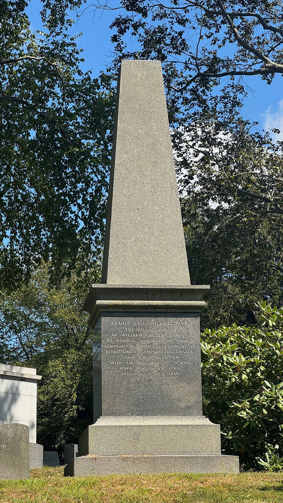 A tall gray stone obelisk monument in a cemetery, inscribed with a dedication to Fanny Fairchild Bryant, wife of William Cullen Bryant, dates indicating her birth in 1797 and death in 1868, surrounded by trees.