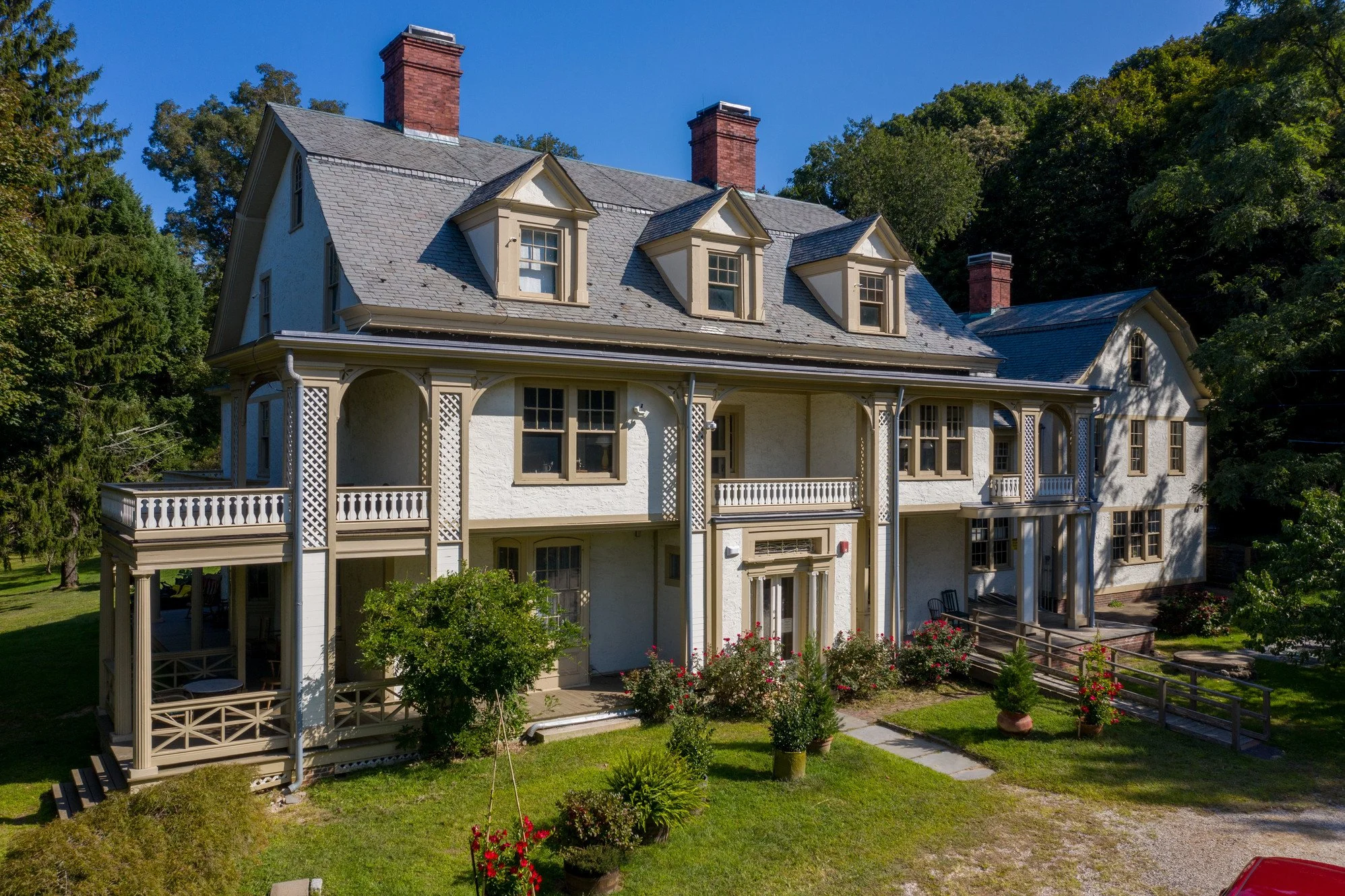 A large white house with multiple levels, a gabled roof, dormer windows, and brick chimneys, surrounded by green trees and a neatly maintained lawn.