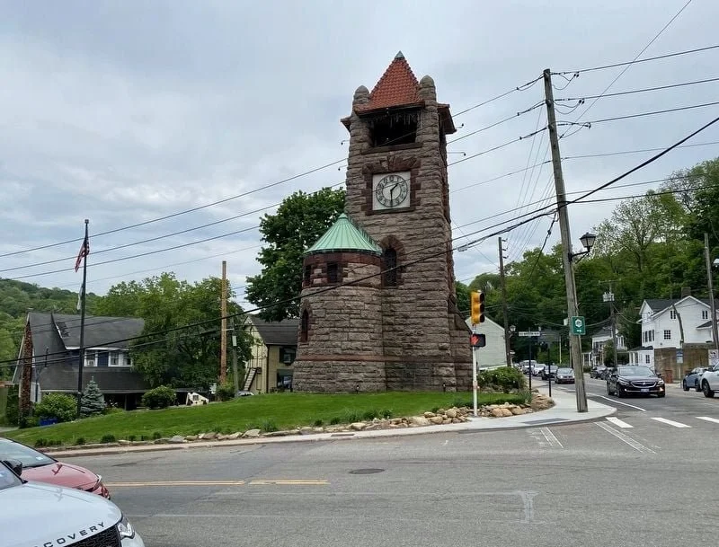 A stone clock tower with a red, pyramid-shaped roof, situated on a grassy area at an intersection, with cars, houses, and trees in the background.