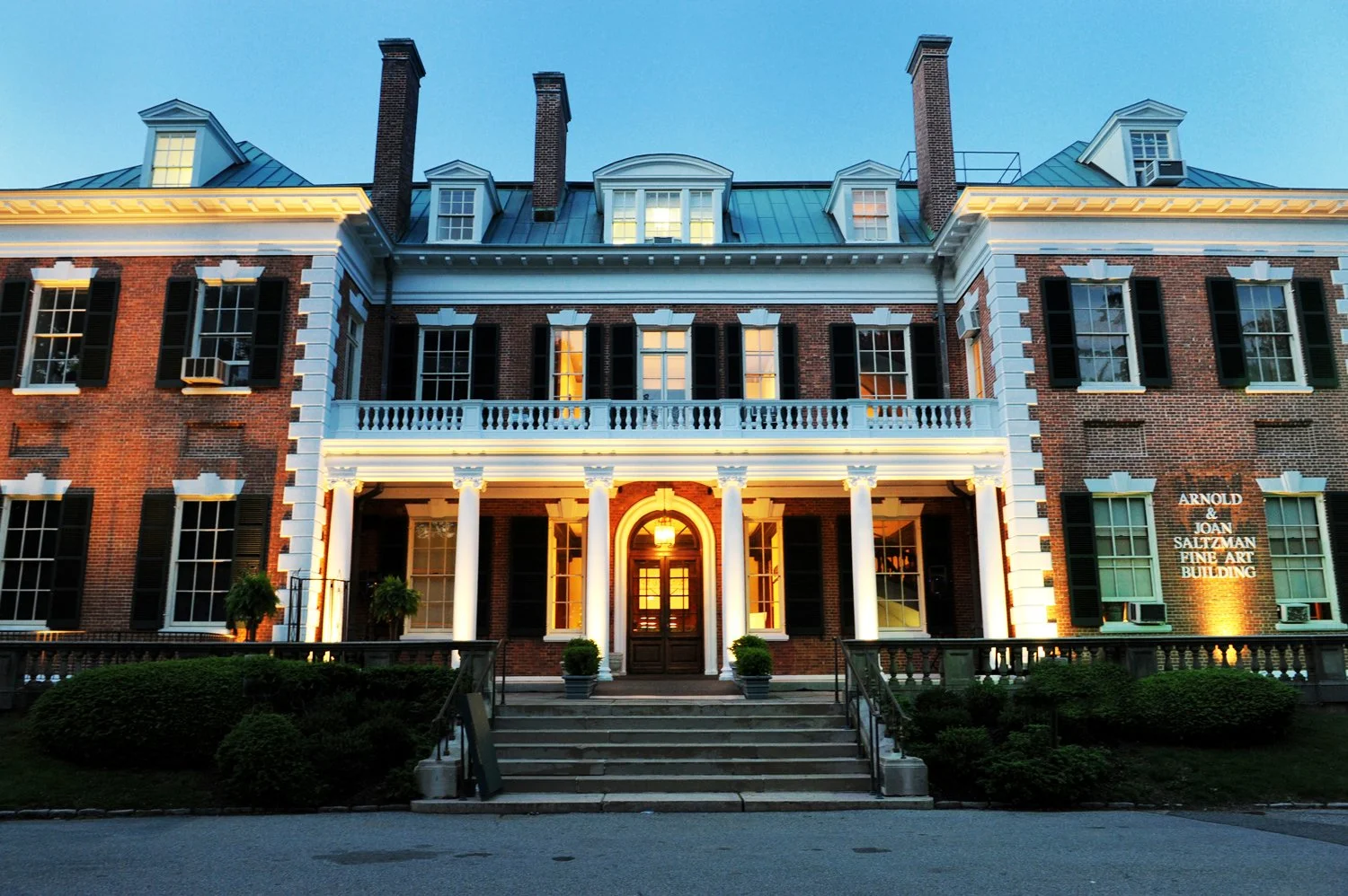 Historic brick building with white columns, black shutters, and a sign reading 'Arnold & Joan Saltzman Fine Art Building' illuminated by exterior lighting at dusk.