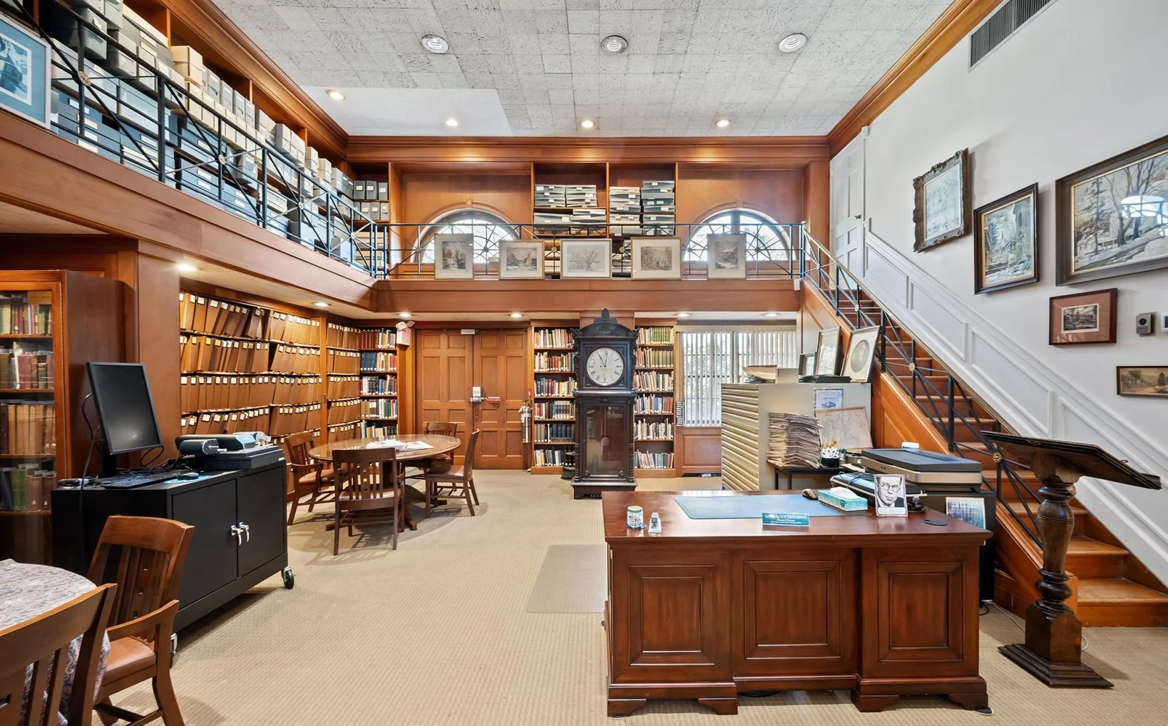 A well-lit library room with wooden shelves filled with books, a large grandfather clock, framed artwork on the walls, a staircase leading to an upper level with more books, and a desk with a computer and various papers.